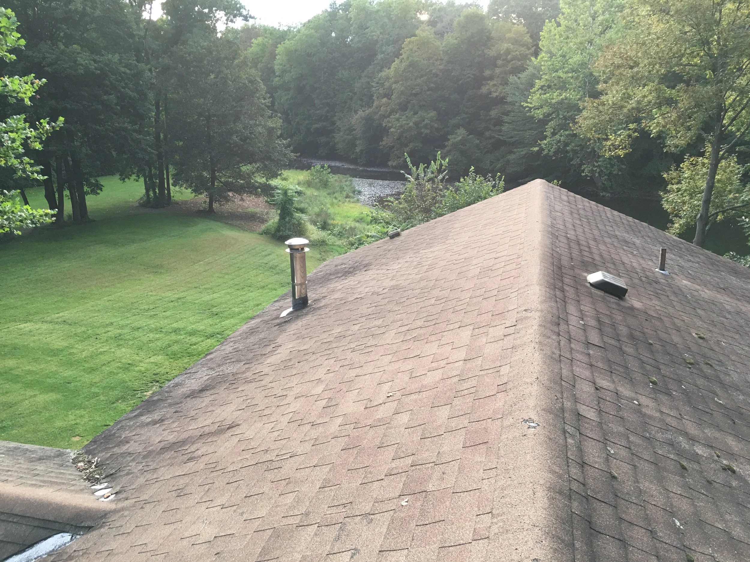 A rooftop with brown shingles, two vents, and some debris. In the background, a green lawn, trees, and a river.