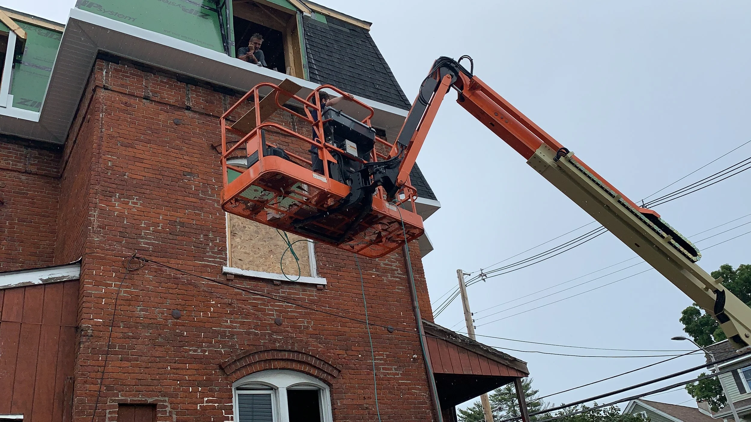 A construction worker climbing in an orange aerial lift basket near an unfinished brick building. The worker is looking at a phone or device, and the building has a partially completed exterior with some windows and siding. Overhead power lines are visible against a cloudy sky.
