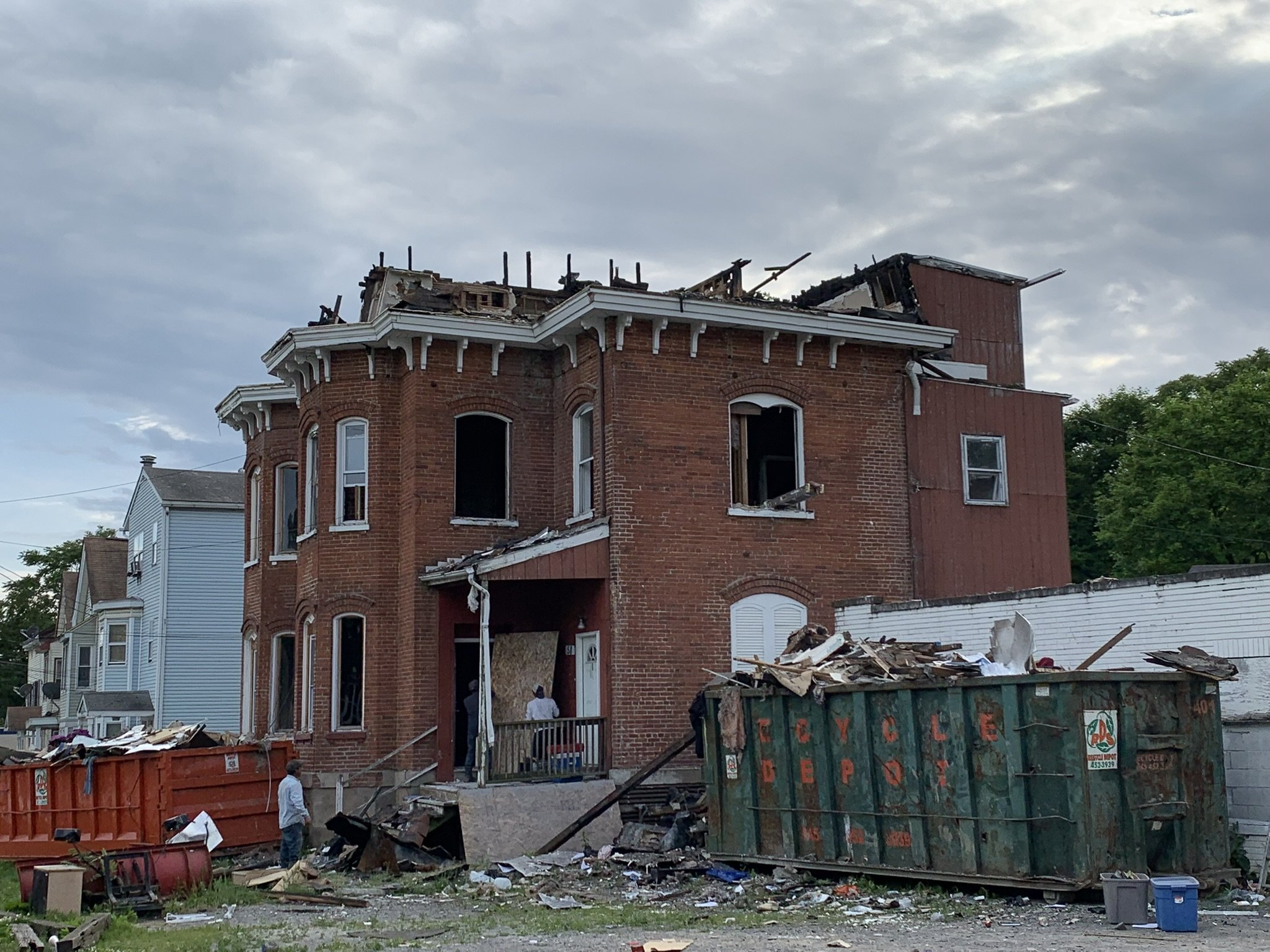 A damaged brick building with part of the roof collapsed, debris scattered in front, and dumpsters filled with debris in a residential neighborhood.