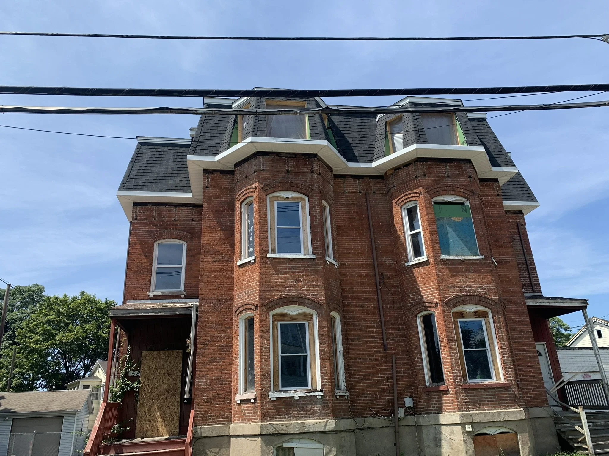 A tall, three-story brick house with ornate bay windows, some with white trim, and a steep, dark shingled roof with multiple gables. The house is under construction or renovation, with boarded-up windows and scaffolding on the side. Power lines are visible in the foreground, and the sky is clear and blue.