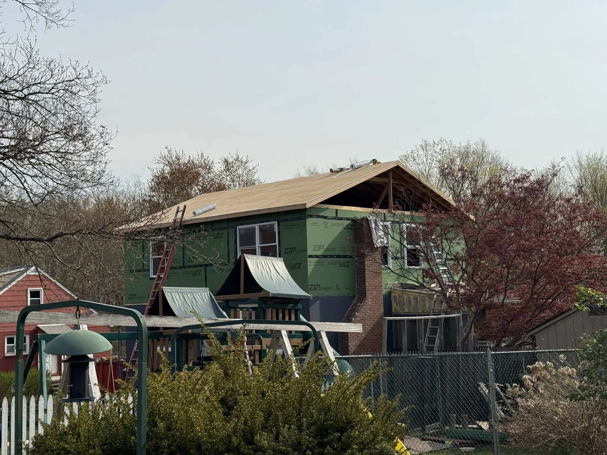 A house under construction with a new roof, surrounded by trees and a backyard with a swing set, trampoline, and fencing.