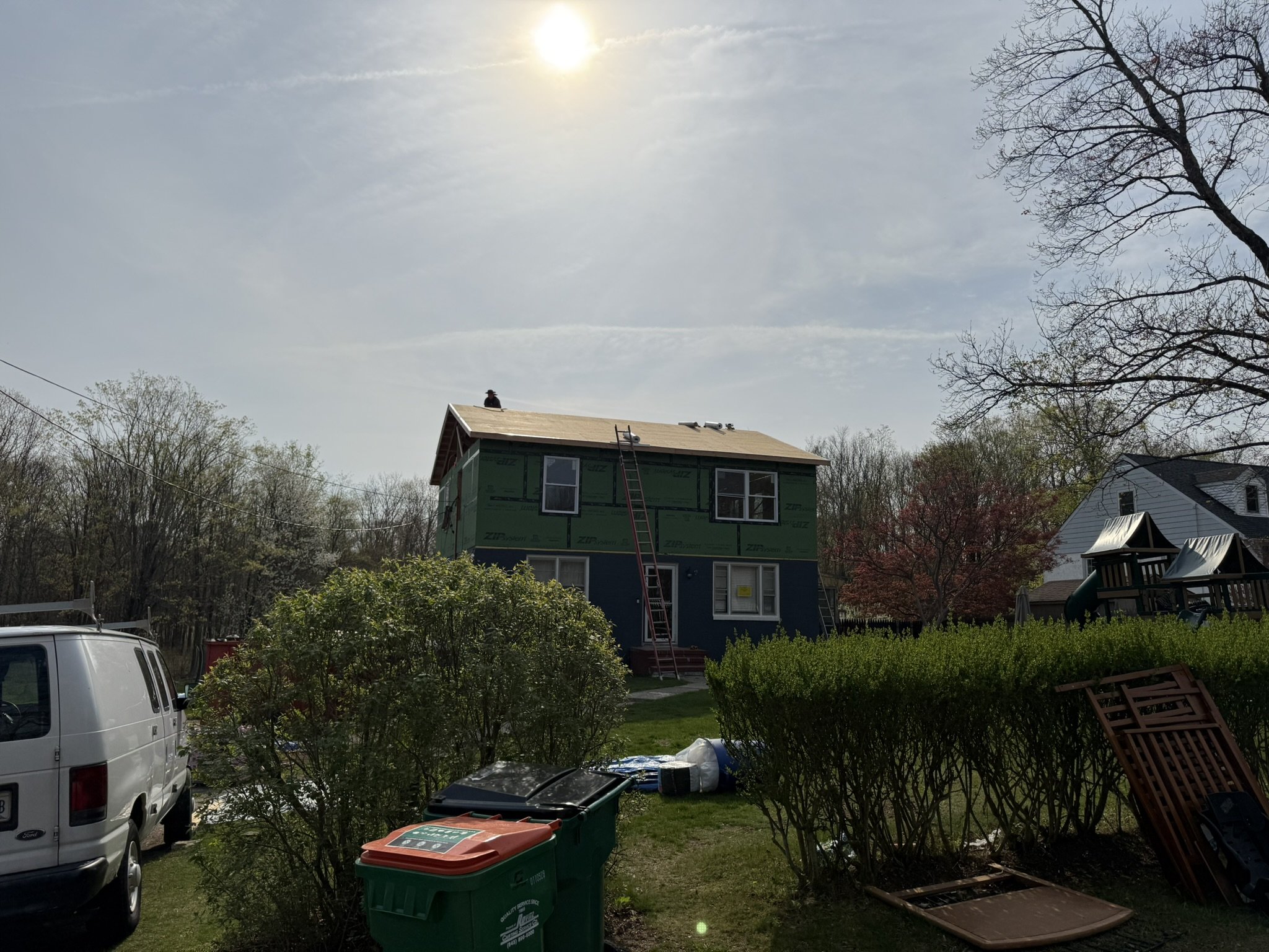 A house under construction with a person on the roof, surrounded by trees and a clear sky with the sun shining.