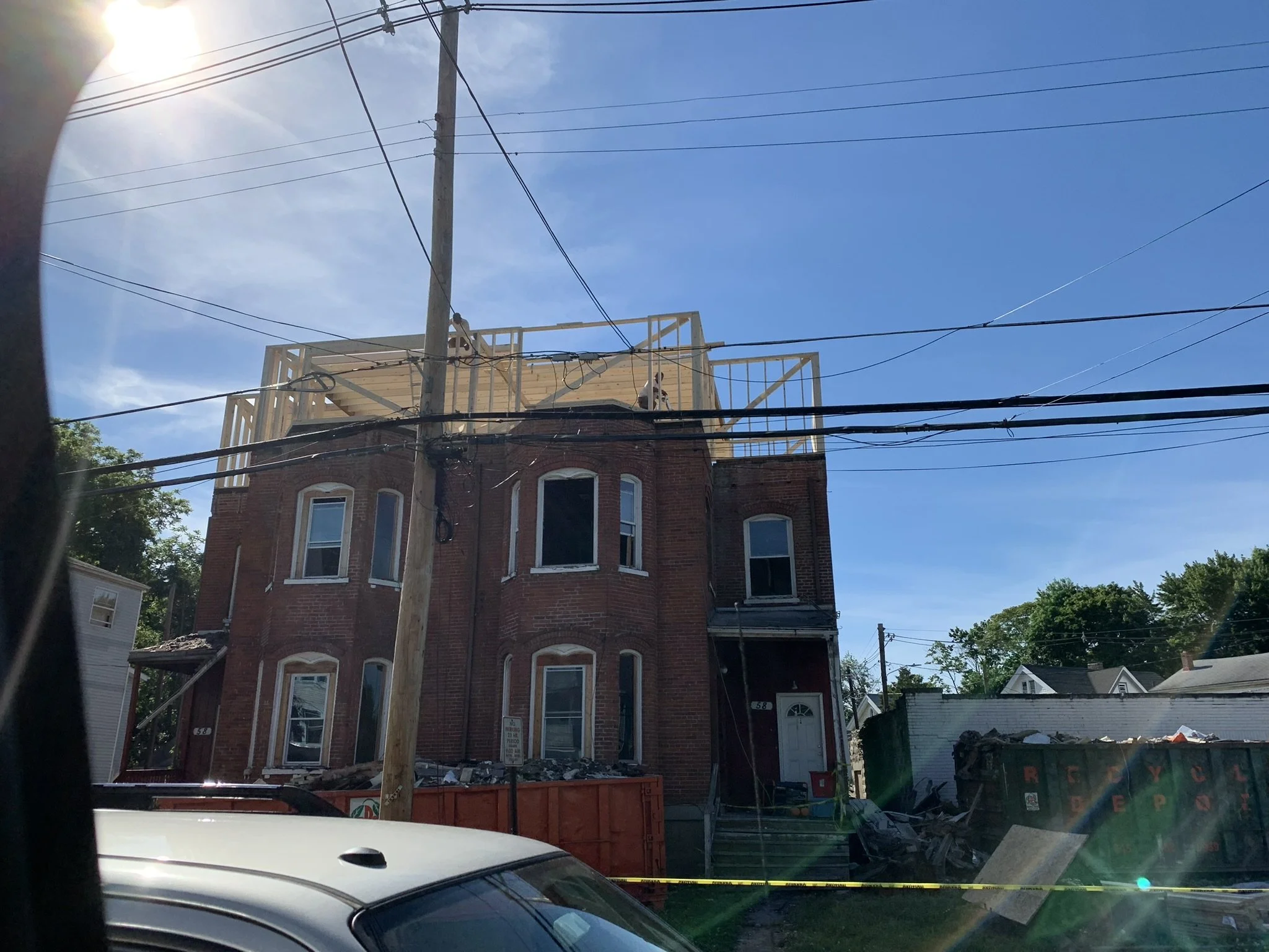 A multi-story brick building under construction with a wooden framework on the top floor. There are power lines crossing in front and a utility pole nearby. Construction materials and debris are visible near the building, and a parking sign is on the street in front.