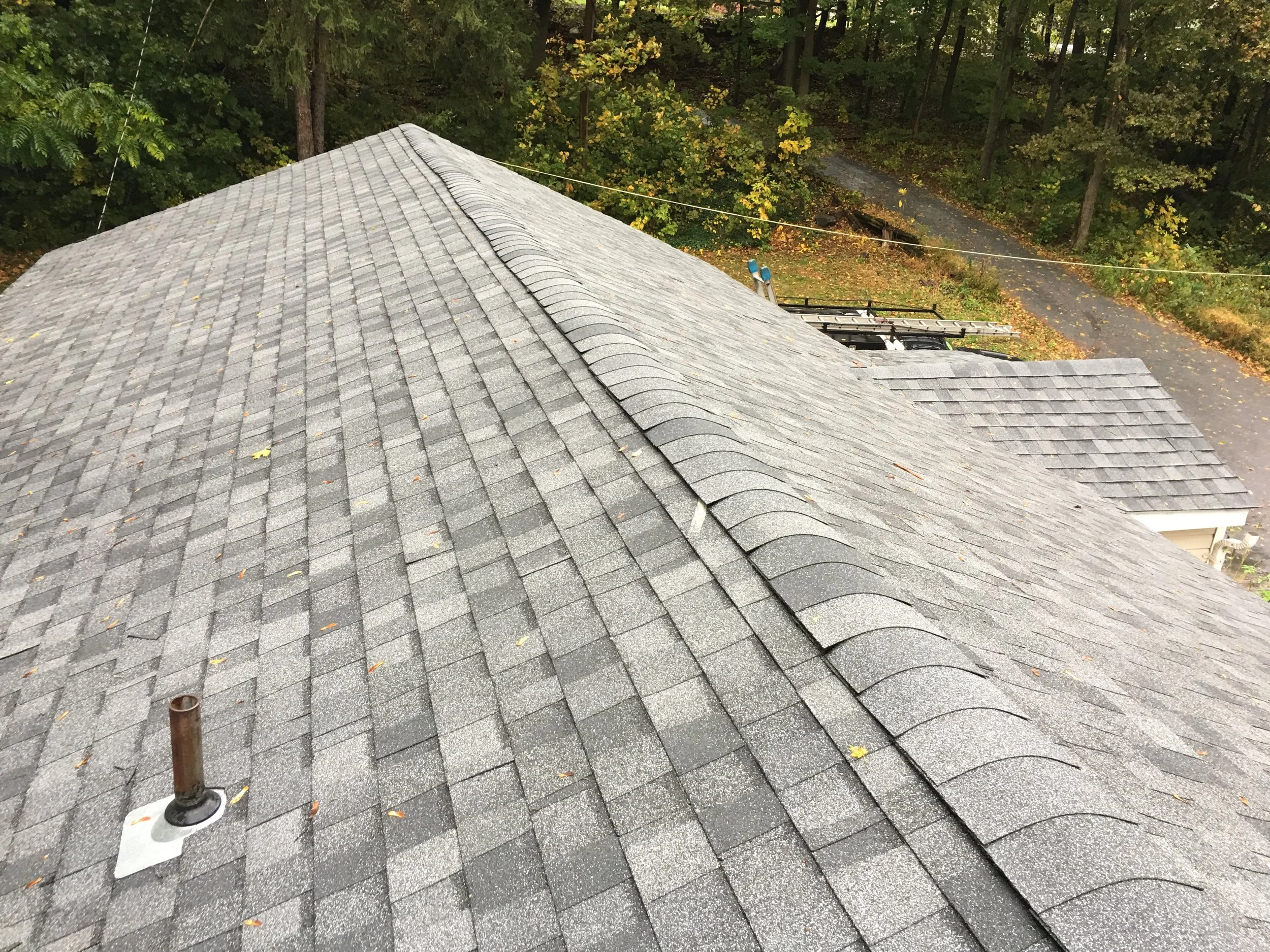 View from a rooftop showing gray shingles, a ladder, a ladder and a cable, with a wooded area and a gravel driveway below, during autumn.