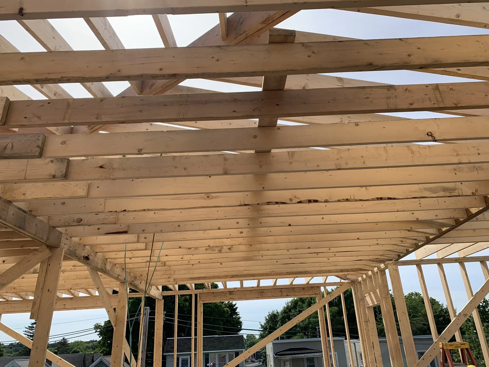 Wooden framing structure under construction, with visible beams and support posts, set against a background of trees and a partly cloudy sky.