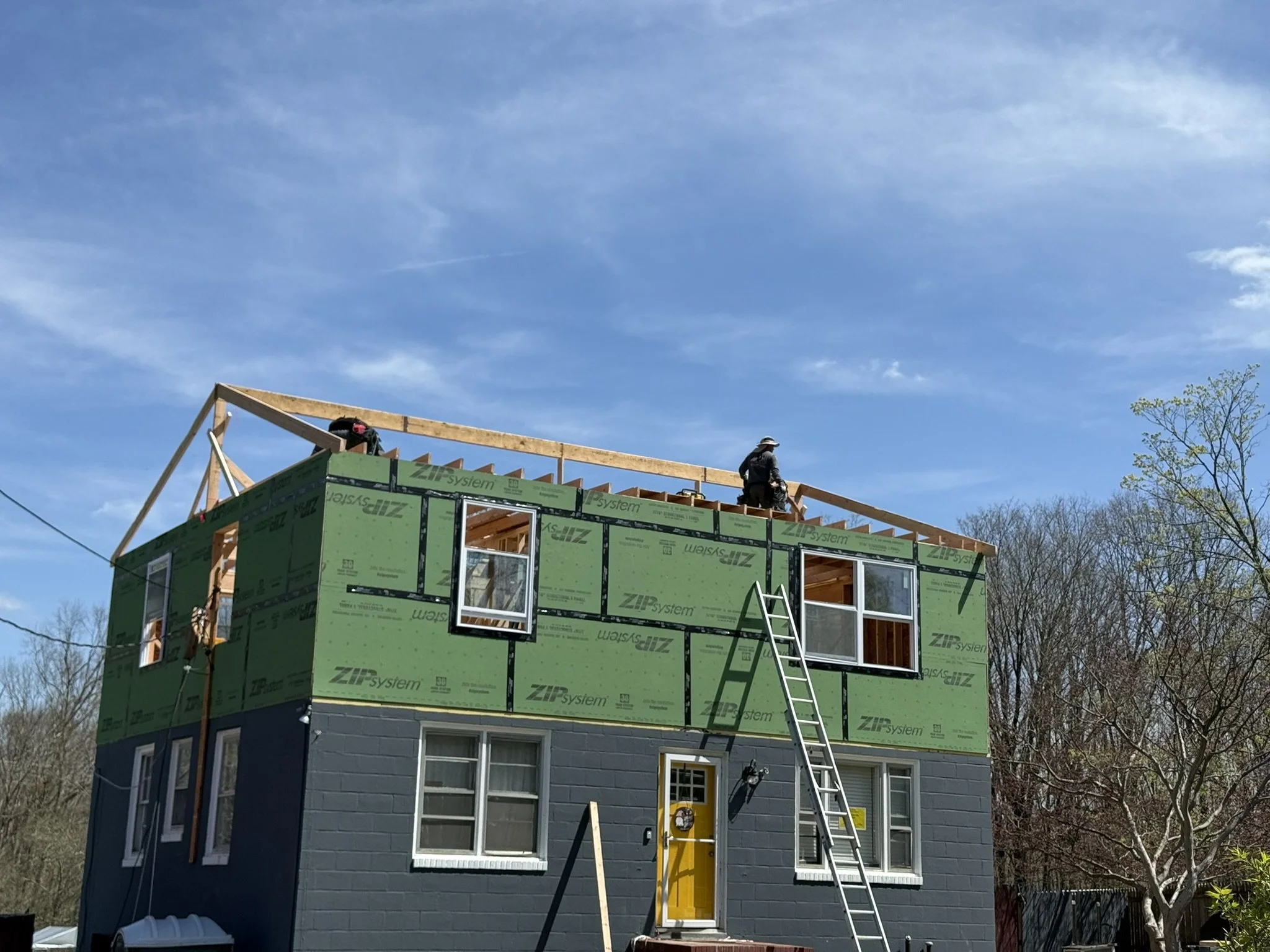 A two-story house under construction with the exterior walls partially covered in green weather-resistant sheathing. The upper part of the house shows framework for a roof, with a worker standing on the roof framing. There is a ladder leaning against the house for access. The house has multiple windows, some of which are installed, and the lower story has a dark-colored wall with additional windows and a bright yellow door. The background features blue sky and bare trees.