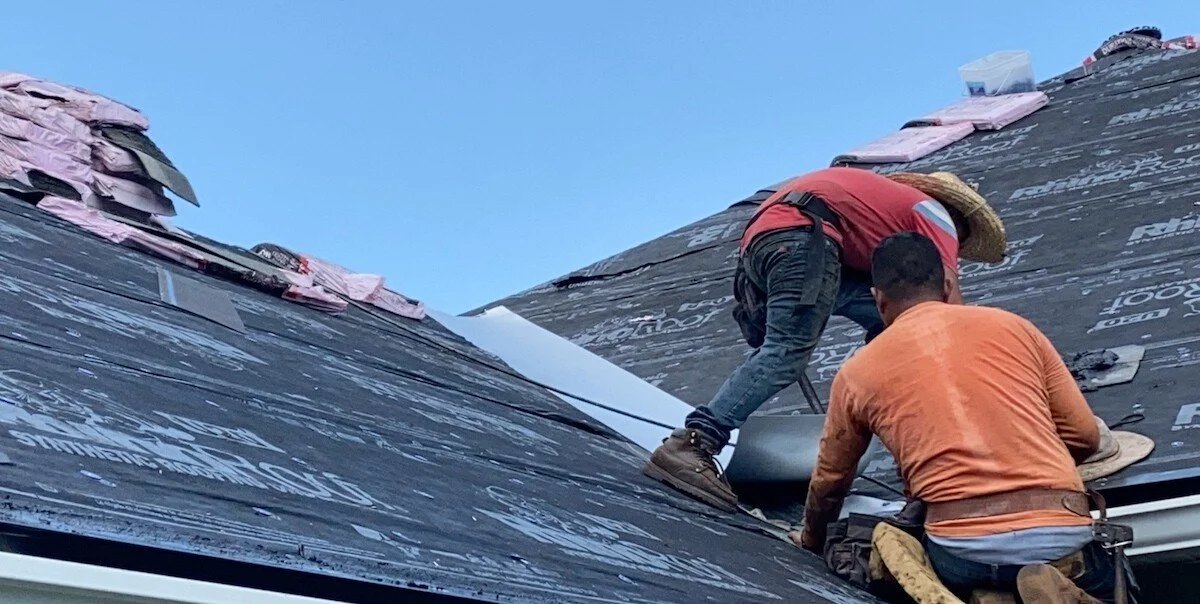 Two workers installing roofing underlayment on a sloped roof during daylight.