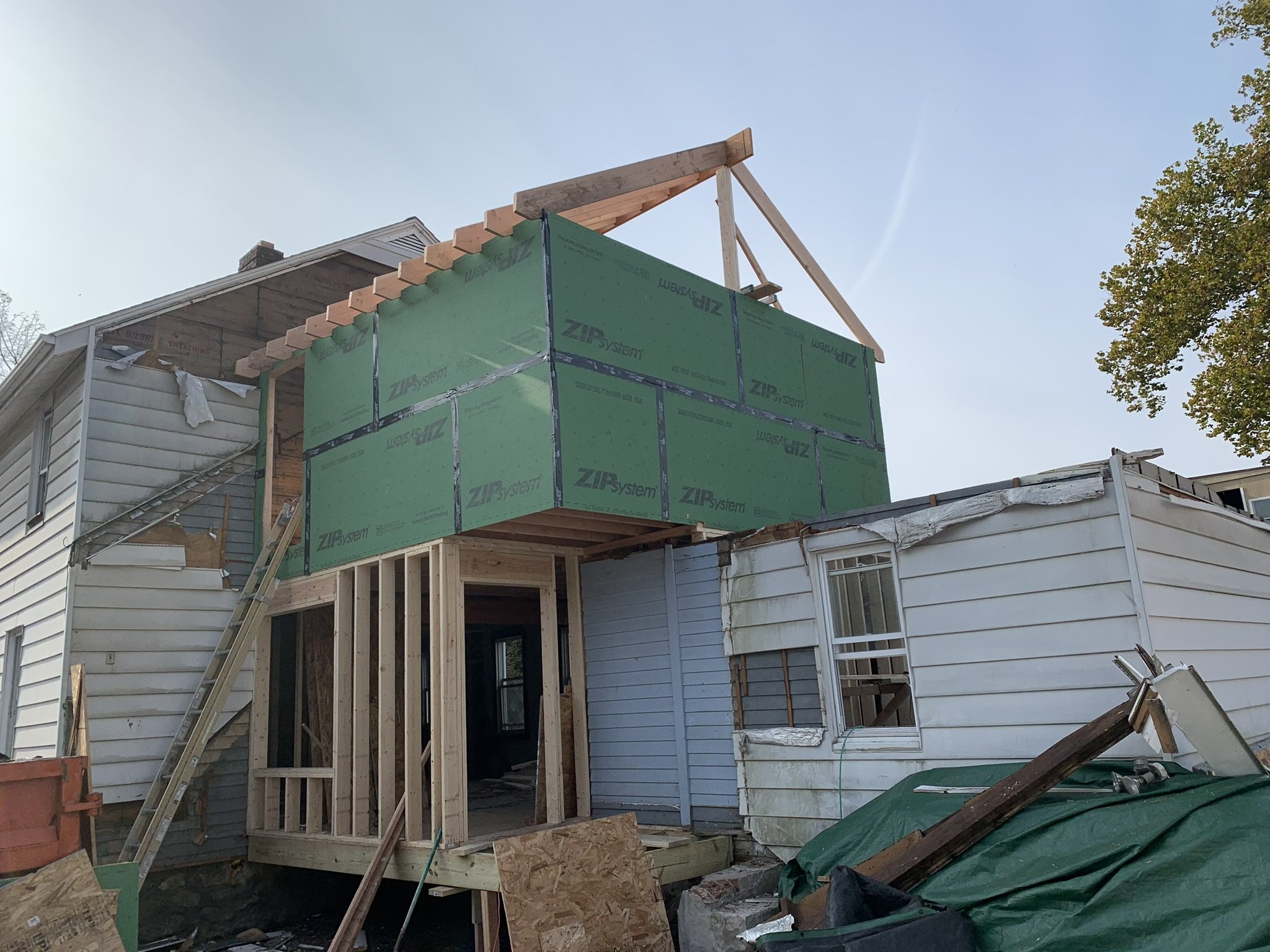 Construction site of a house with framing and green insulation or sheathing, partial roof work, and surrounding building materials and debris.