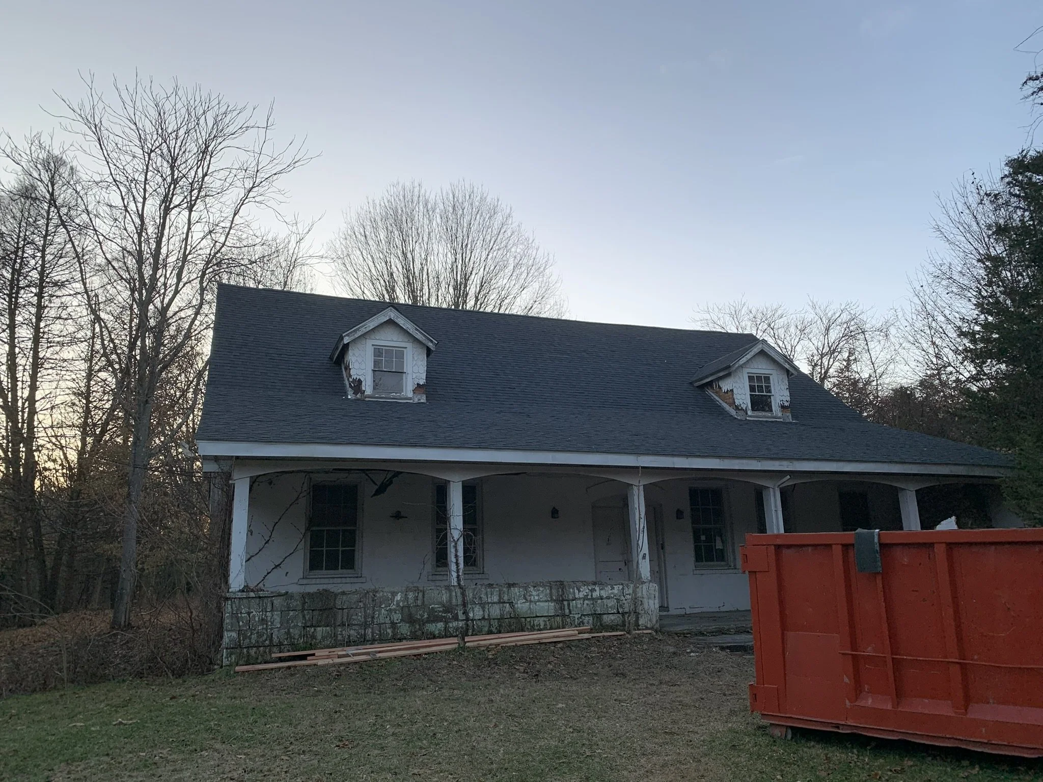 Backyard view of a house with a gray roof and two small dormer windows. Trees without leaves are visible in the background, and a red dumpster is in the foreground on the right side. The sky is clear with a subtle blue gradient.