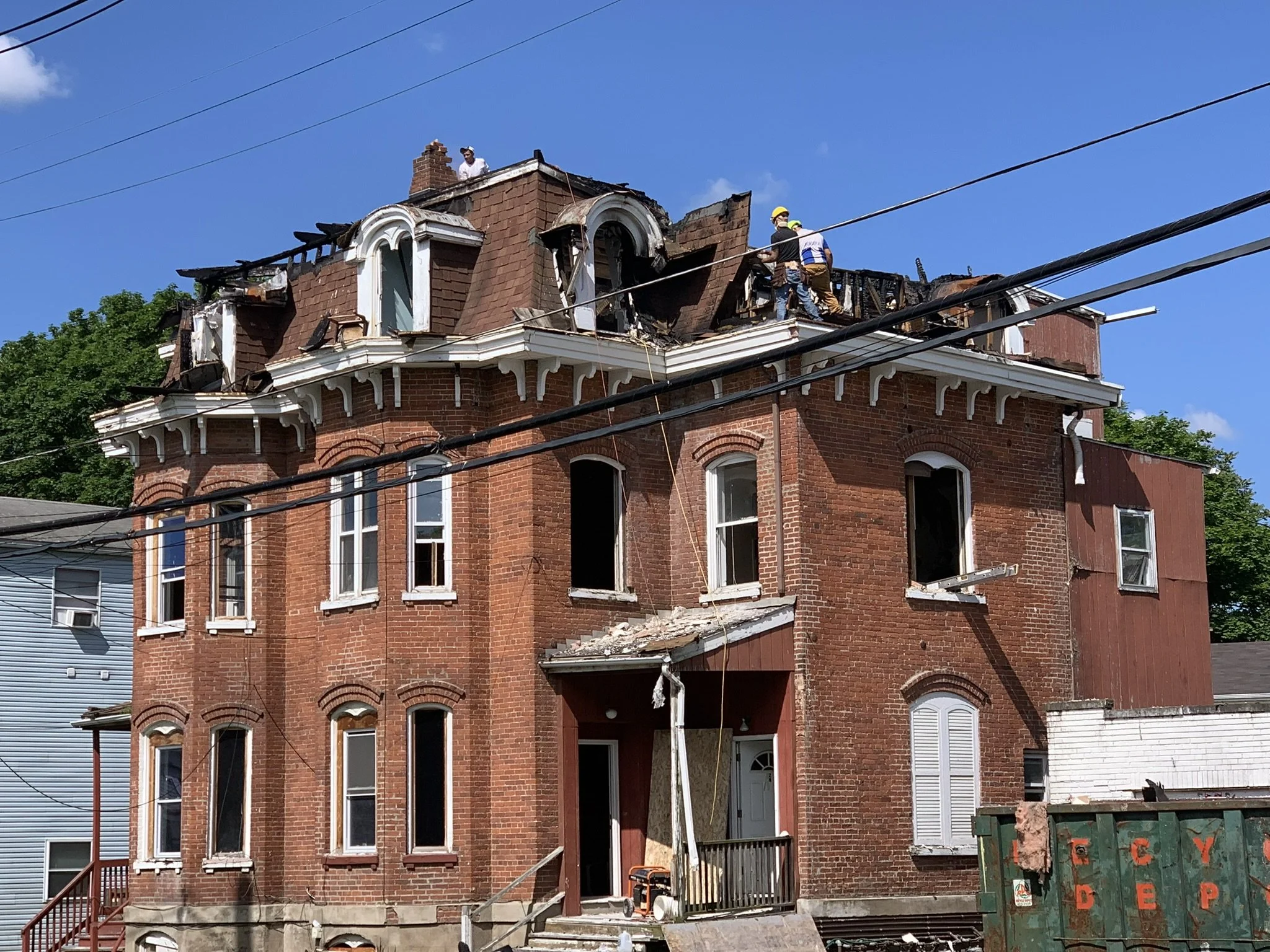 A three-story brick house with extensive fire damage on the upper floor, with charred and missing sections of the roof and walls, and several workers wearing safety helmets on the roof inspecting the damage.