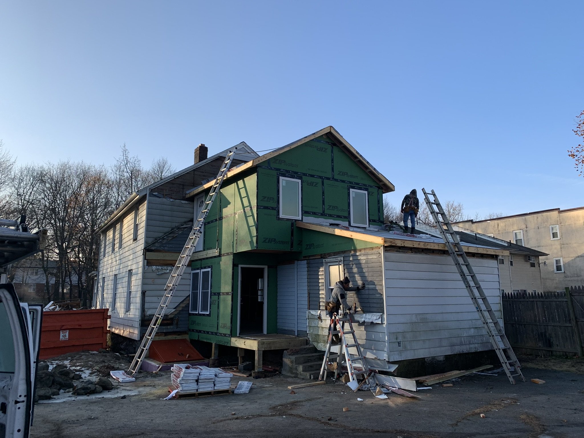 A house under construction or renovation with workers and ladders on the roof. The house is partly covered with green insulation material, and there are building materials and tools on the ground.