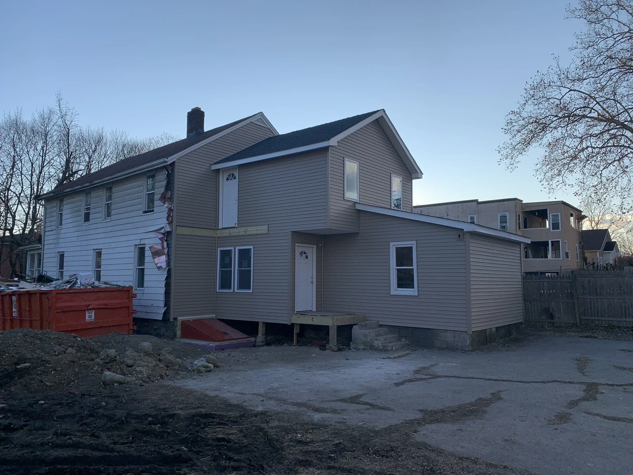 A house under construction with beige siding, some windows, and a white door, with the yard still being prepared and a construction dumpster in the front yard.