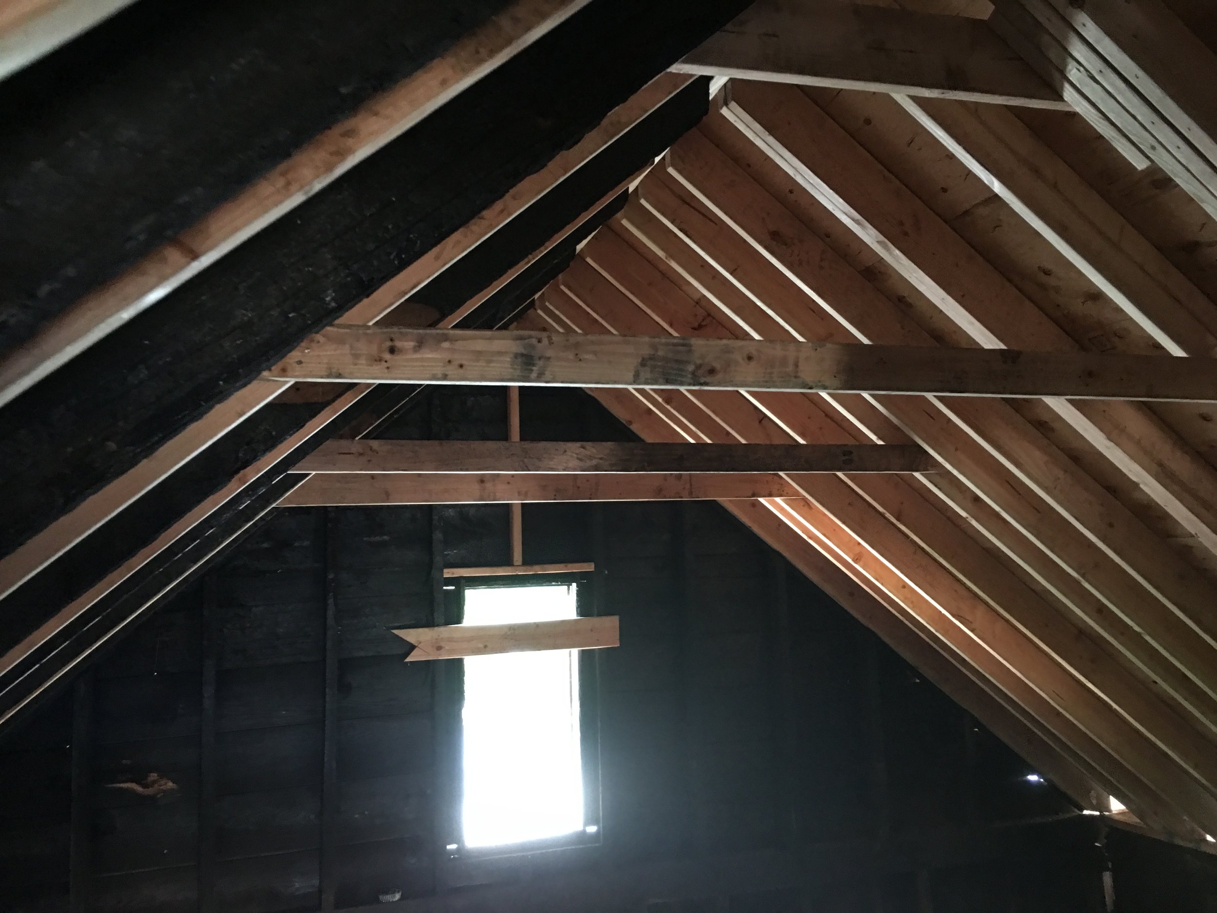 Interior view of an unfinished attic with wooden roof rafters and a large opening at the end, allowing natural light to enter.