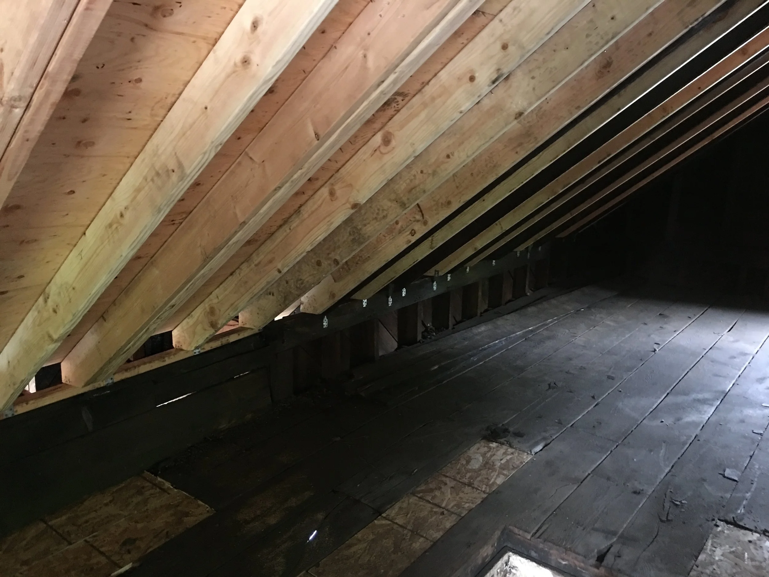 Underside view of a slanted wooden roof with visible rafters and unfinished flooring, dark area beneath.
