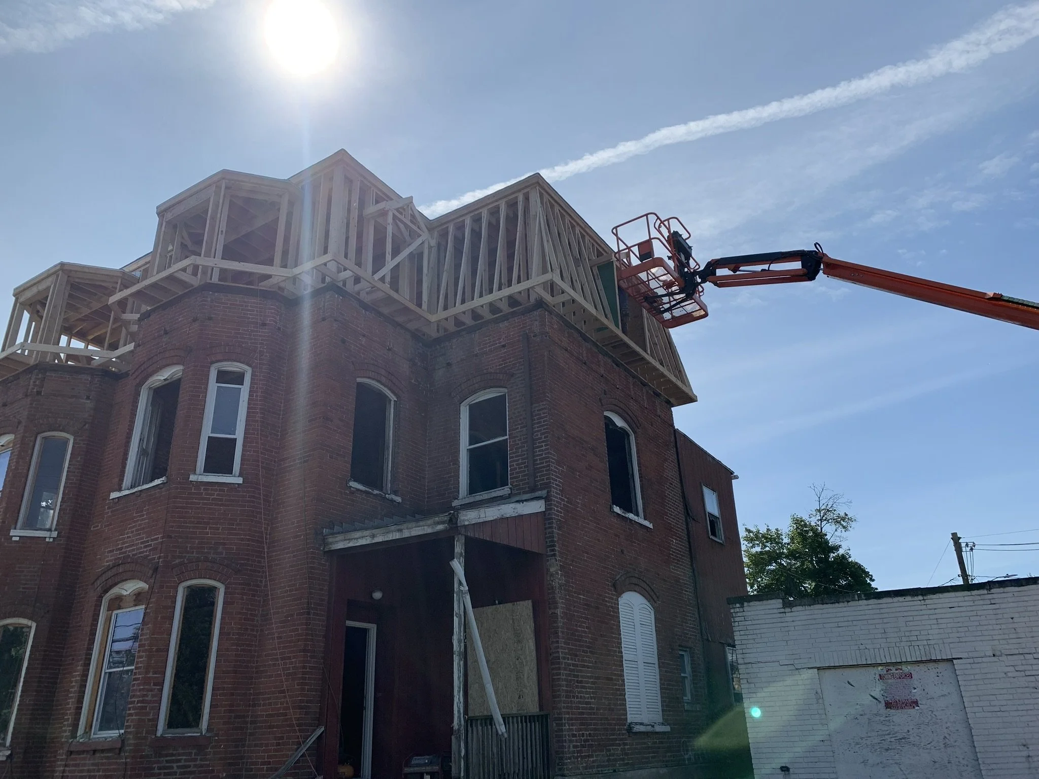 A multi-story brick building under construction with wooden framing on the upper levels, a cherry picker lift extending to the top, bright sunlight, and a clear blue sky with some wispy clouds.