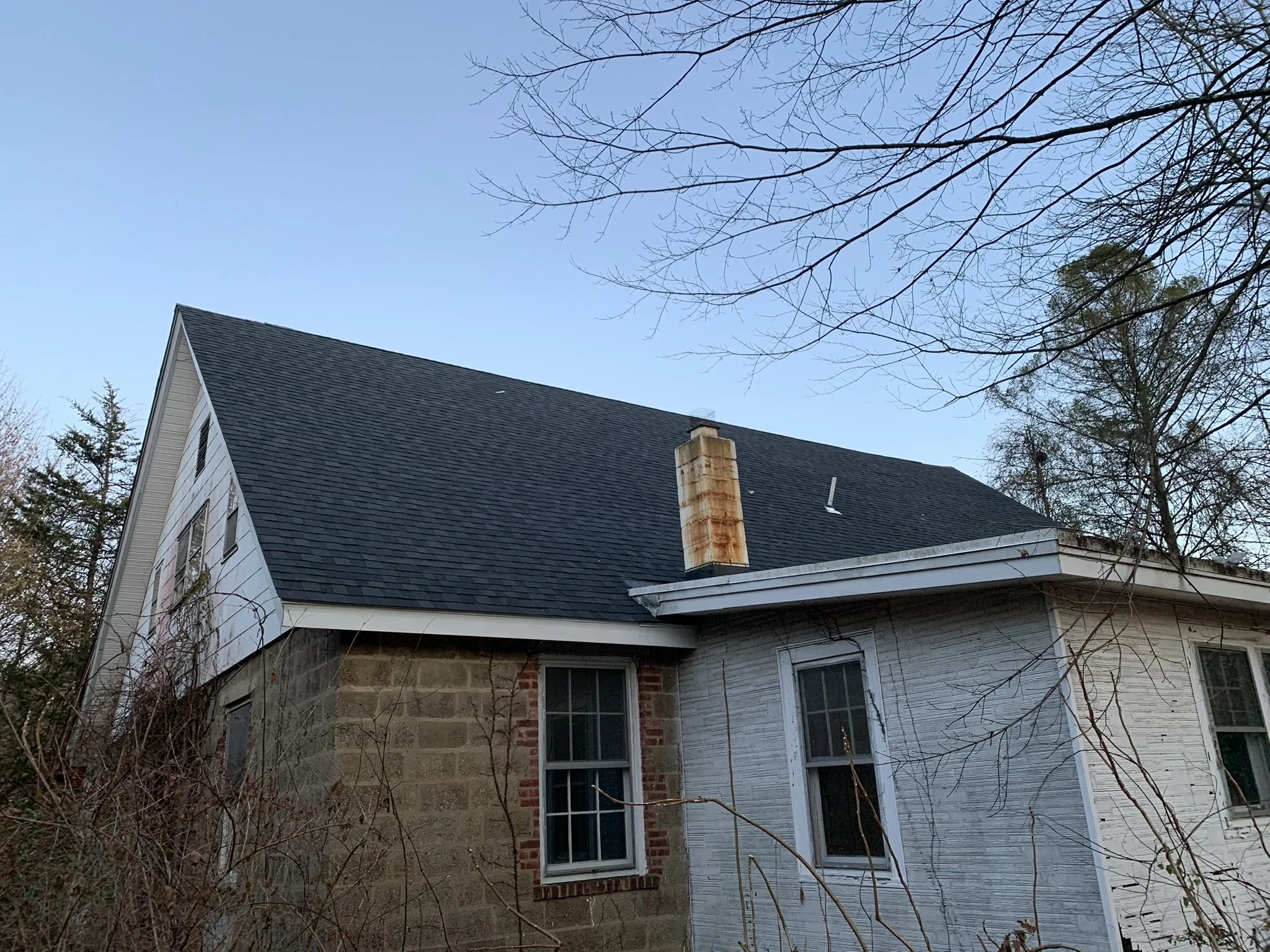 A house with a dark gray sloped roof, white siding, and window frames. The house has a partially brick and stone exterior at the lower level. Trees with leafless branches are in the foreground and background, under a clear blue sky.