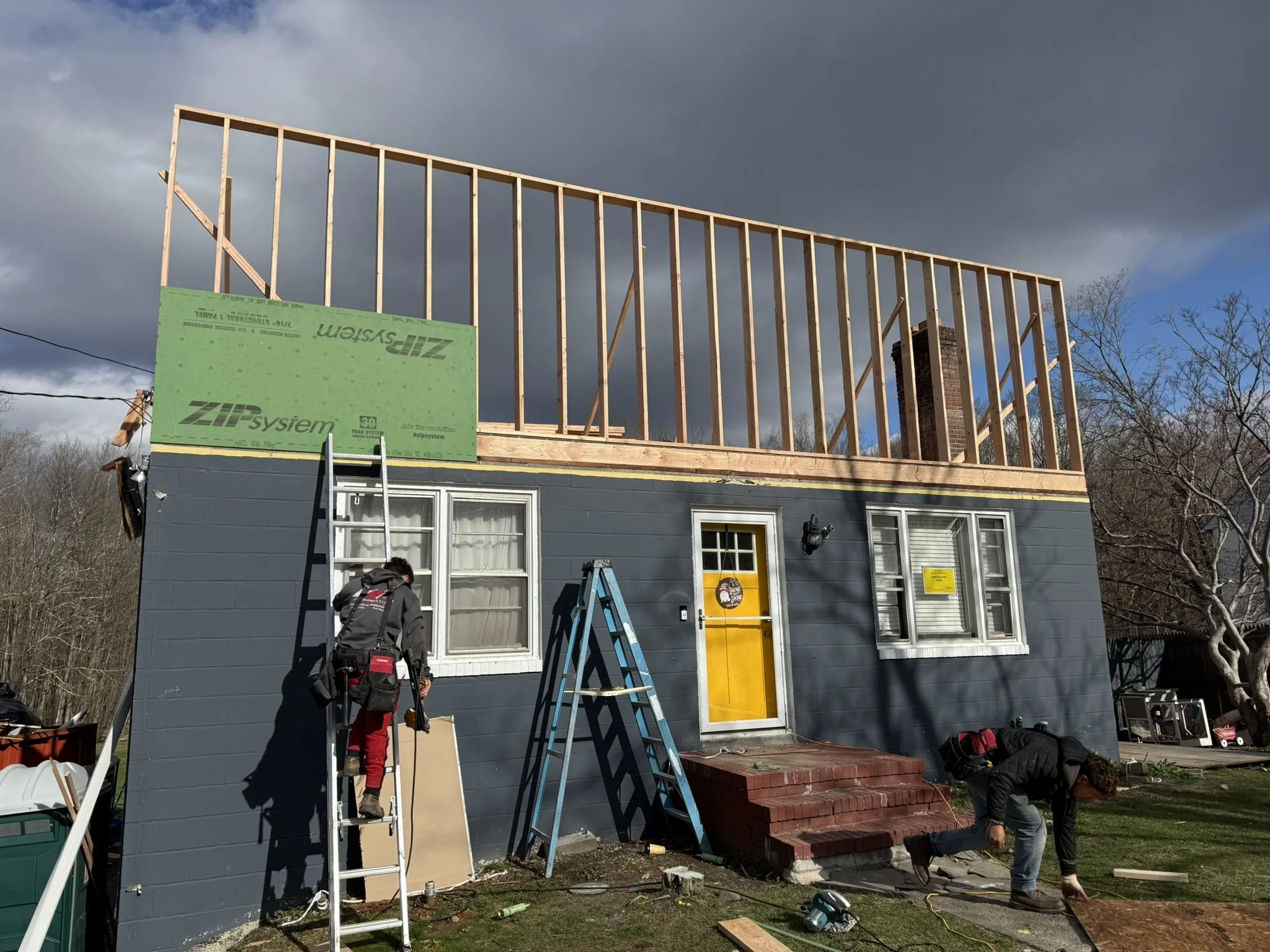 Construction workers building the upper floor of a house with wooden framing, ladder, and brick stairs, under cloudy sky.