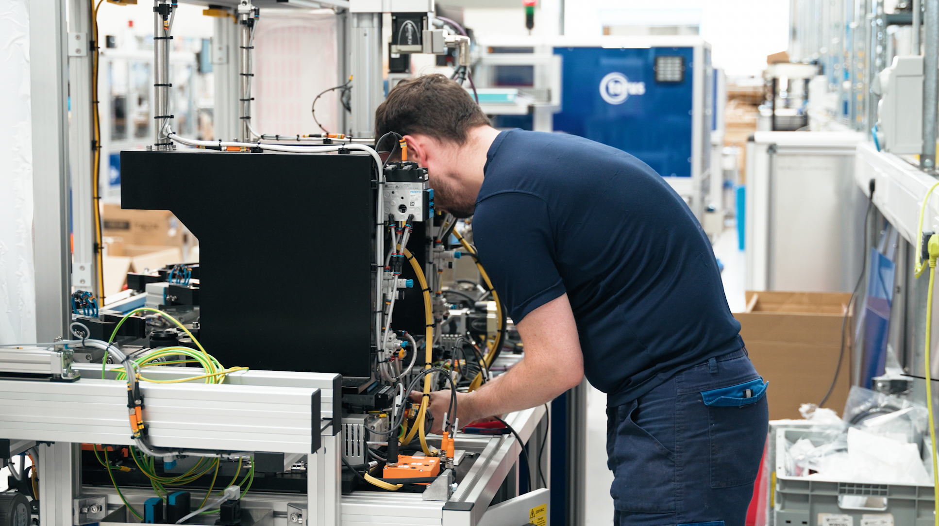 Engineer working on an intricate electronic or robotic device, closely inspecting or assembling it in a well-lit industrial or laboratory environment, surrounded by machinery and wiring.