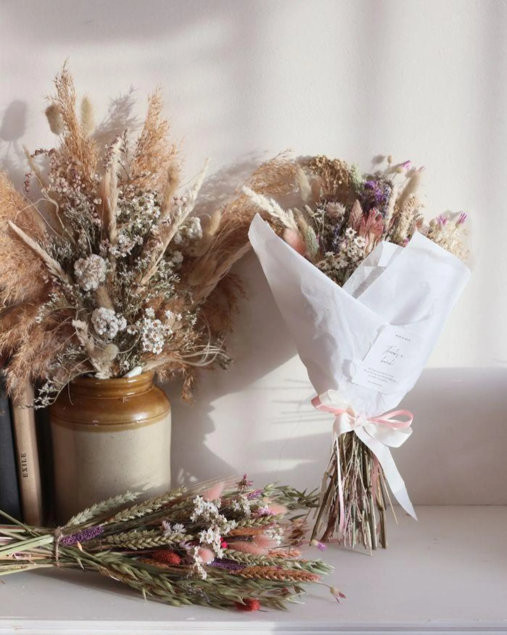 A bouquet of dried flowers and grasses wrapped in white paper, tied with a pink ribbon, leaning against a wall above a ceramic vase with a branch of dried lavender in a well-lit room.