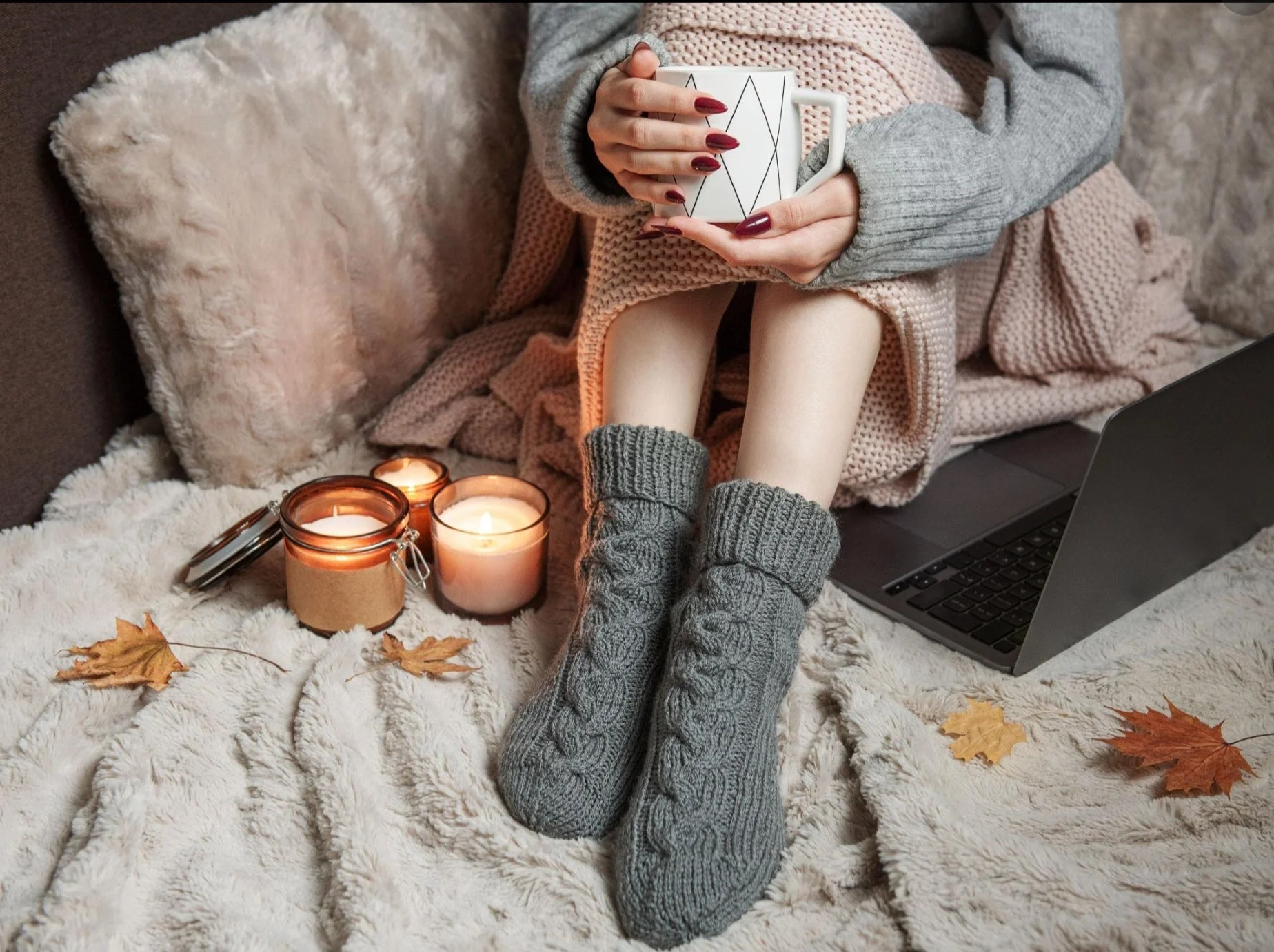 Person sitting on a cozy, furry rug with candles, a laptop, and autumn leaves, holding a mug with geometric patterns, dressed in warm, gray knit accessories, creating a warm, autumnal indoor scene.