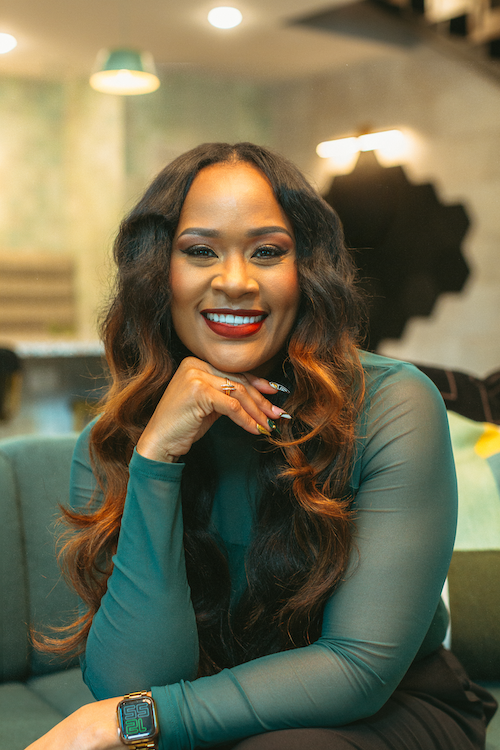 A woman with long, wavy hair and bright red lipstick smiling and resting her chin on her hand in a modern indoor setting.
