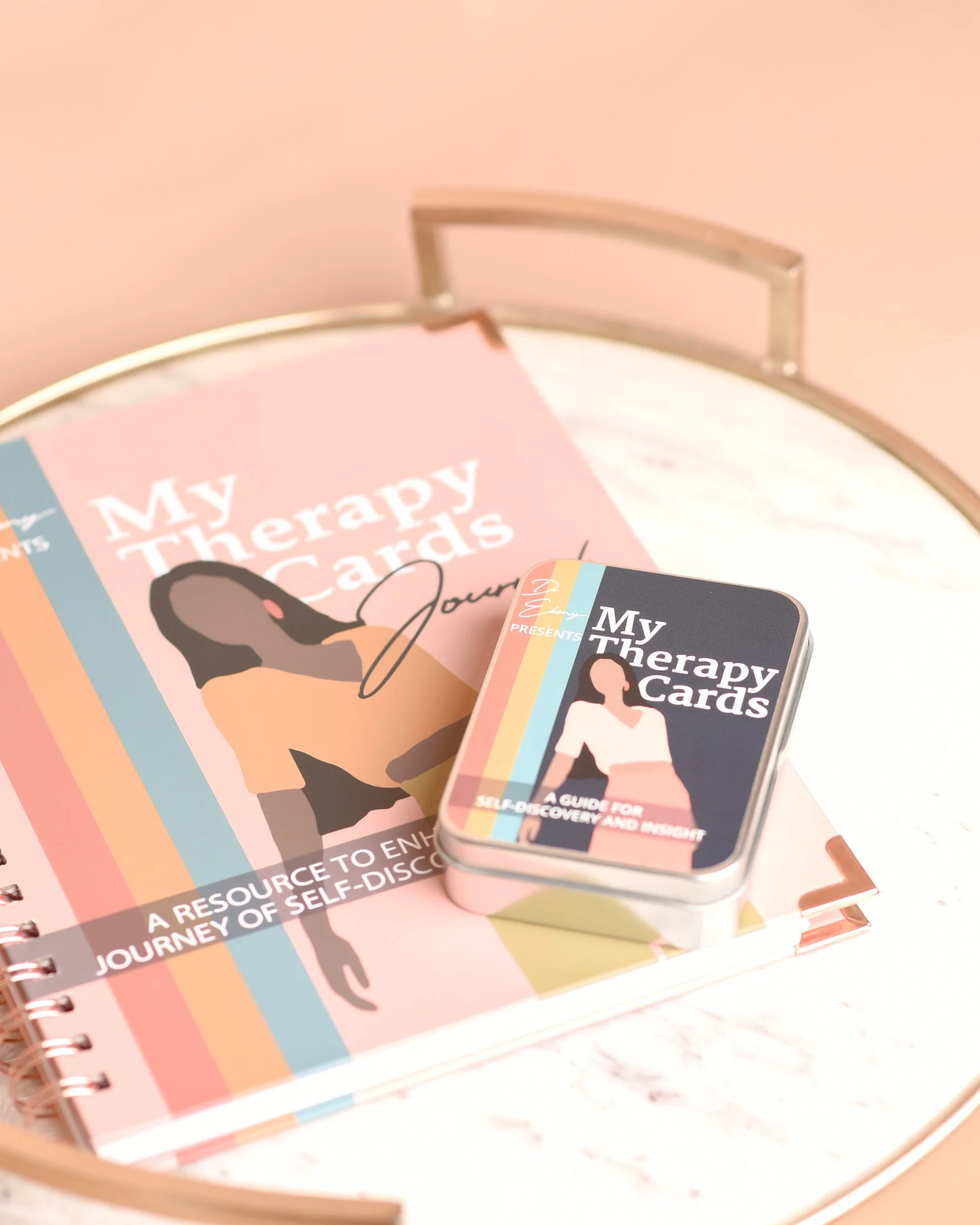 A box of therapy cards titled "My Therapy Cards" placed on a spiral-bound workbook. The workbook cover reads, "A resource to enjoy the journey of self-discovery." The therapy cards box displays a woman in a white top and pink shorts, with her hands on her hips.