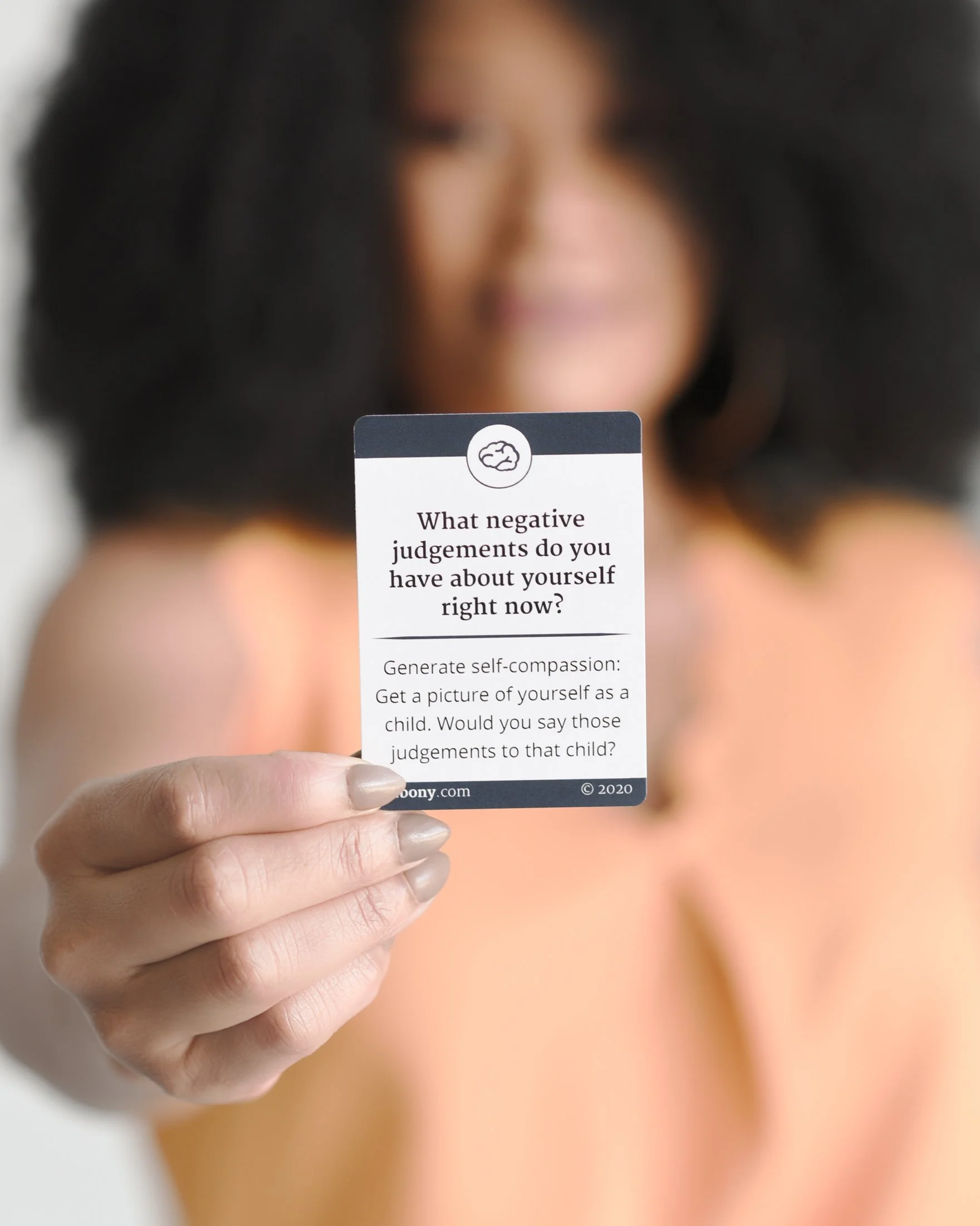 Close-up of a person's hand holding a card with a question about negative judgments and self-compassion, blurred face of a woman with dark hair in the background.