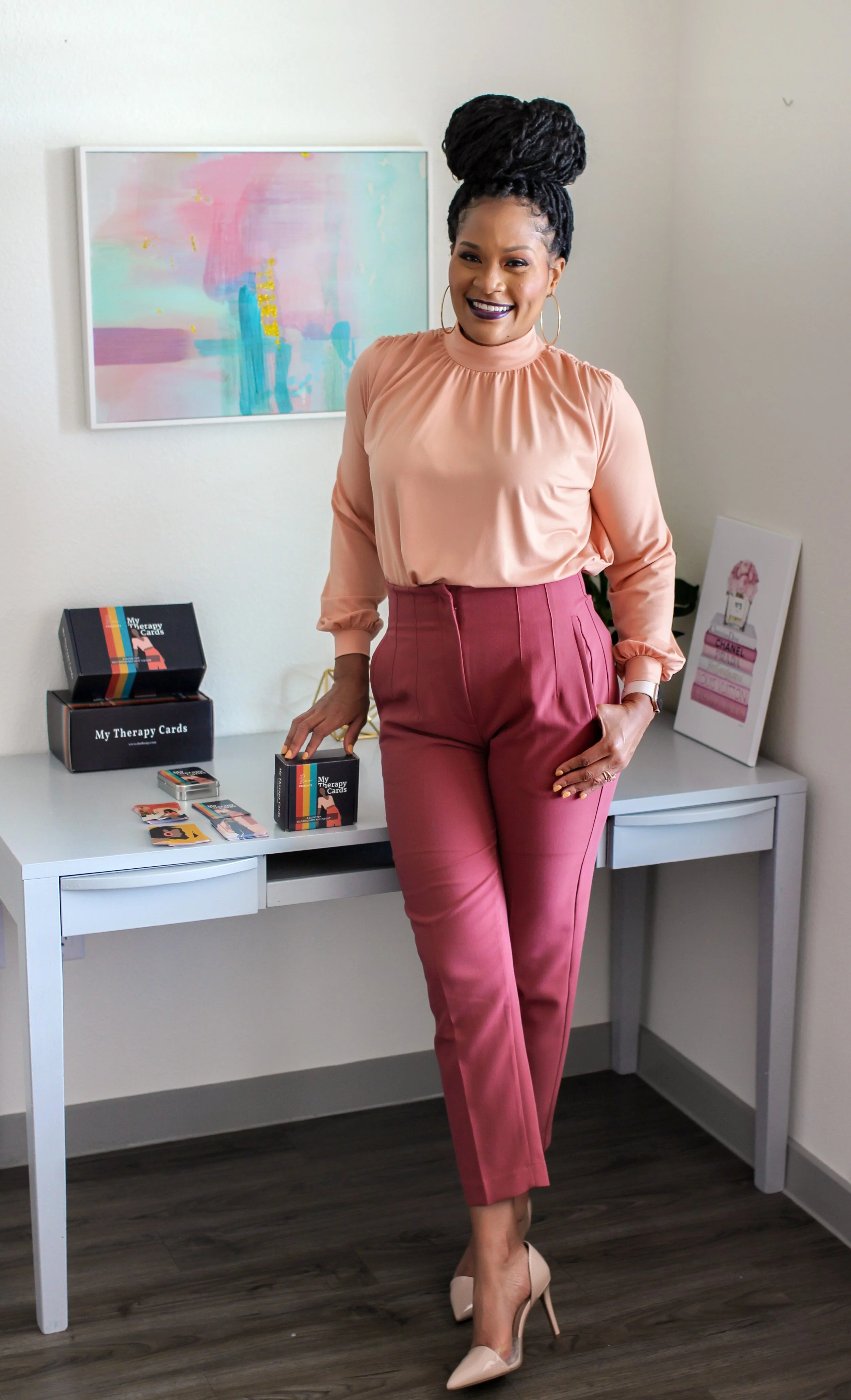 A woman in peach blouse and pink pants standing next to a white desk, smiling. The desk has boxes and cards labeled 'My Therapy Cards.' There is colorful abstract art on the wall behind her.