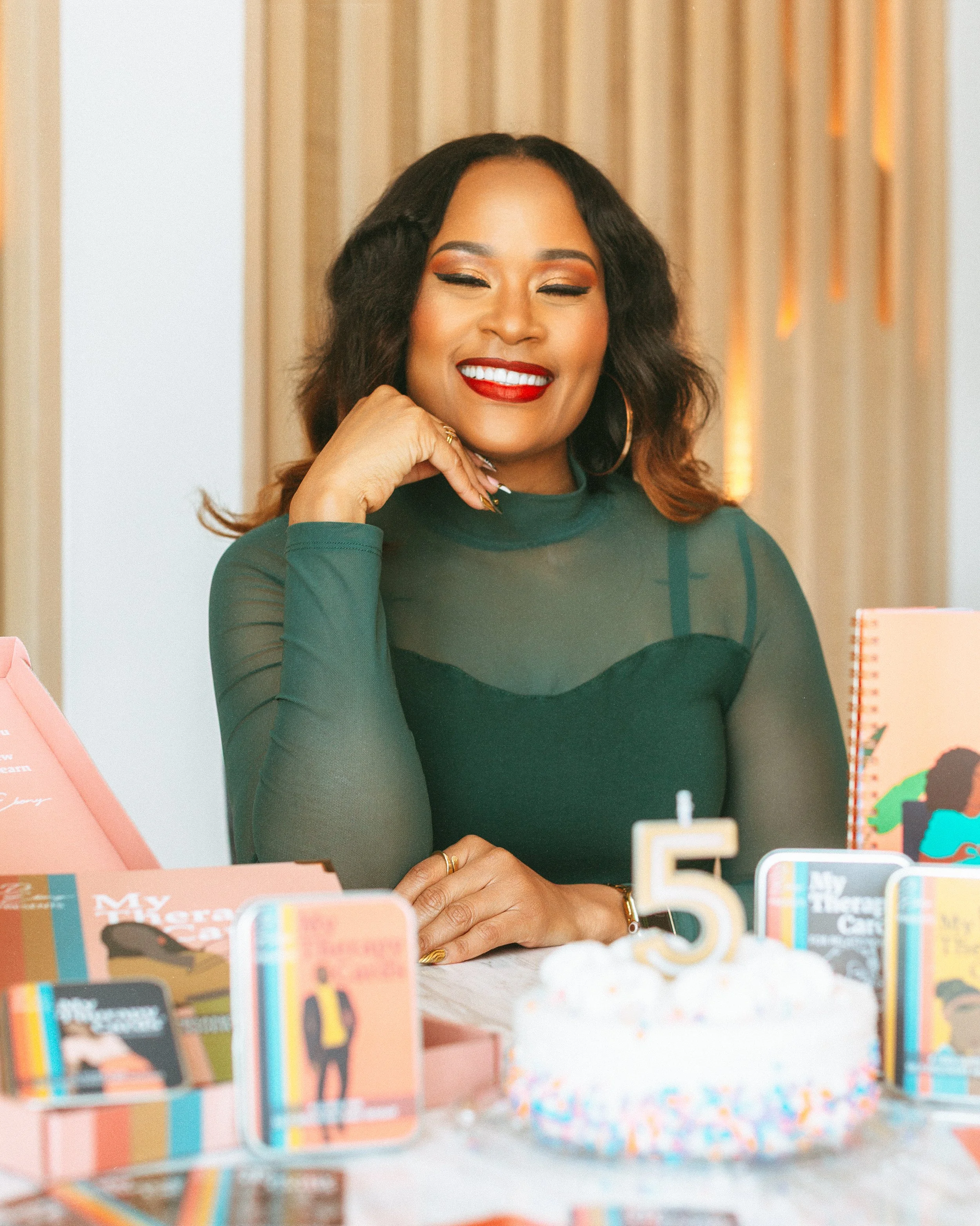 A woman with dark hair, red lipstick, and hoop earrings, smiling with her eyes closed, sitting at a table with birthday decorations and books.