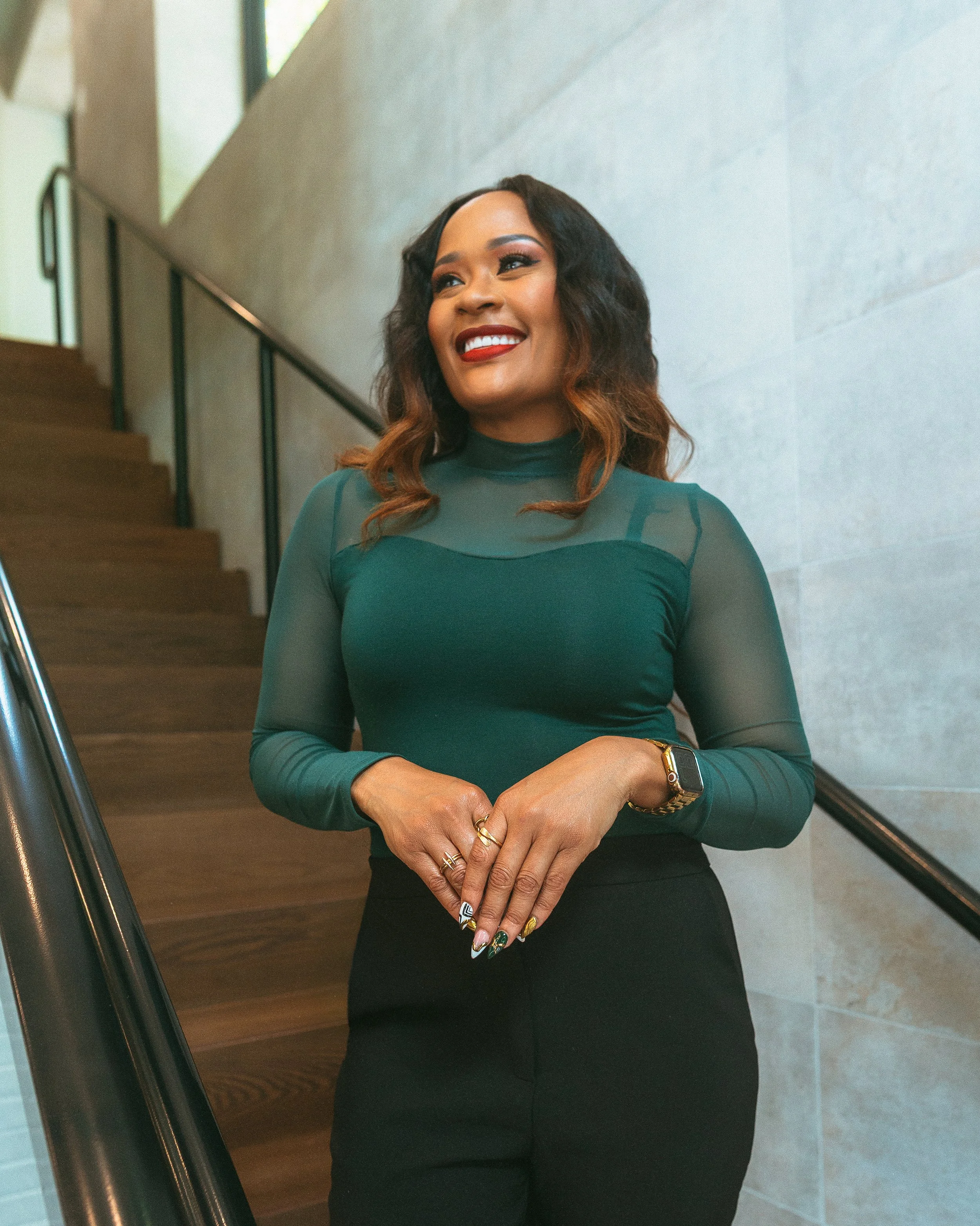 A woman with wavy hair, wearing a dark green sheer long sleeve top and a black skirt, smiling and standing indoors near stairs with a light-colored wall in the background.
