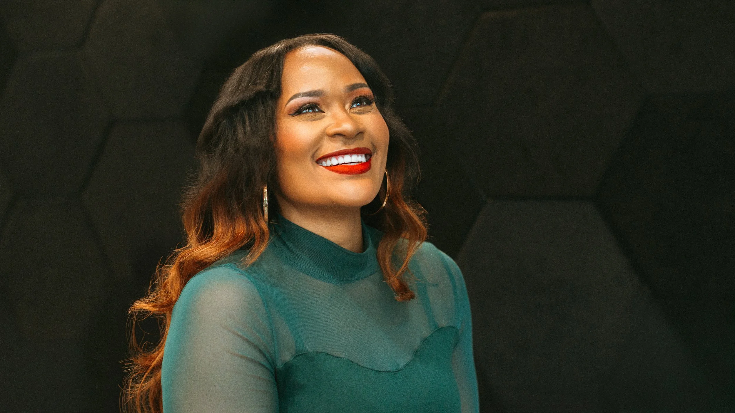 A woman with long wavy hair, wearing a teal top, smiling with red lipstick, earrings, and eyeliner.