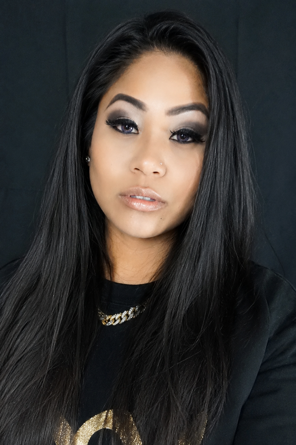 Close-up portrait of a woman with long black hair, wearing makeup, a nose piercing, a black shirt with gold accents, and a gold chain necklace, against a dark background.