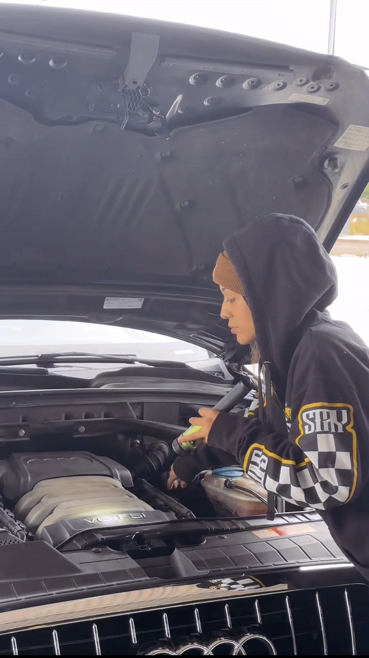 A person wearing a black hoodie with a brown beanie and a black jacket with checkered and yellow patches working on a car engine with the hood open. The person is holding a spray bottle and working inside the engine compartment.