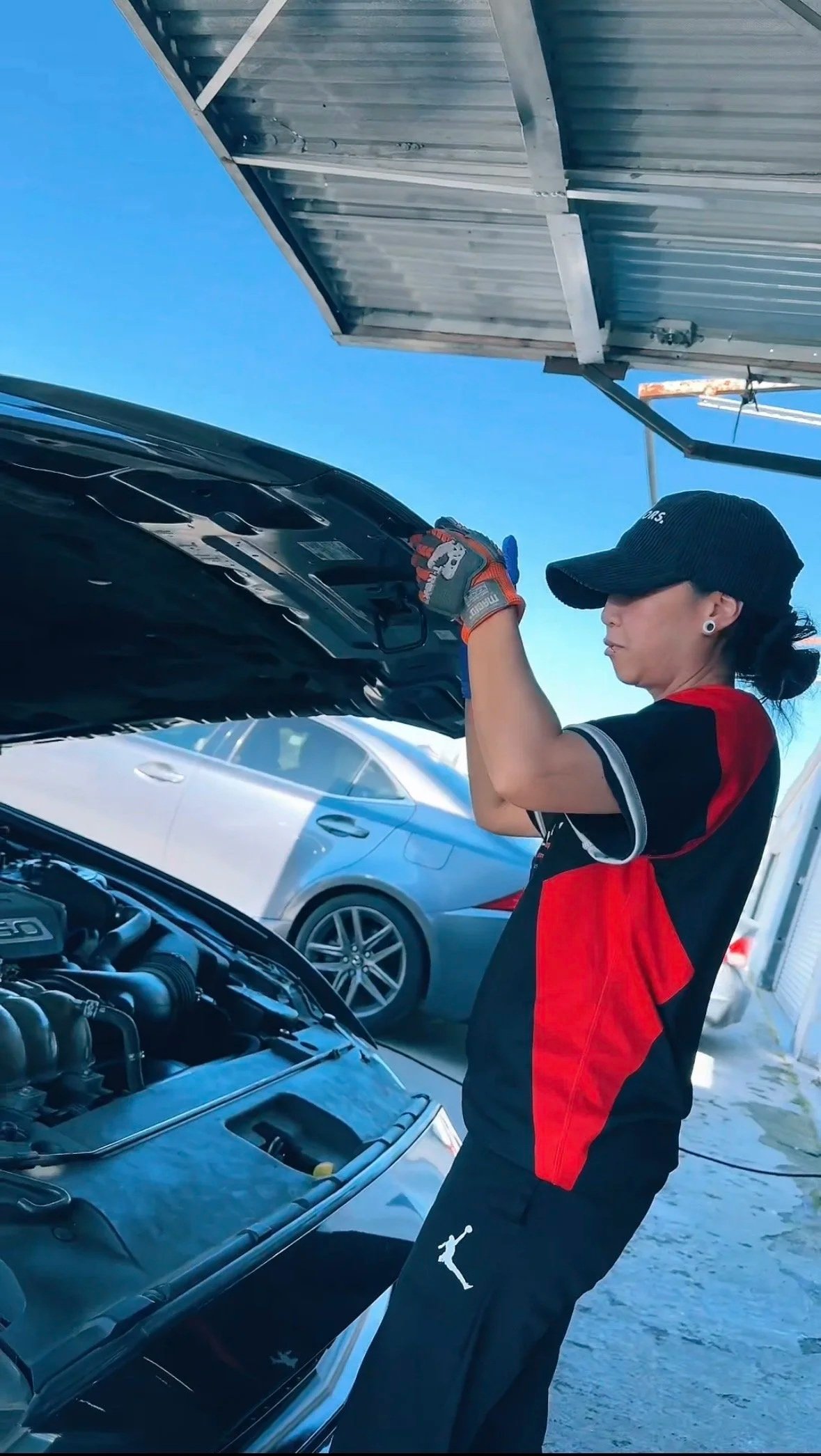 A woman wearing a black cap, a red and black shirt, and black pants with a white logo, inspecting the engine of a car with its hood open at an outdoor location with other vehicles in the background.