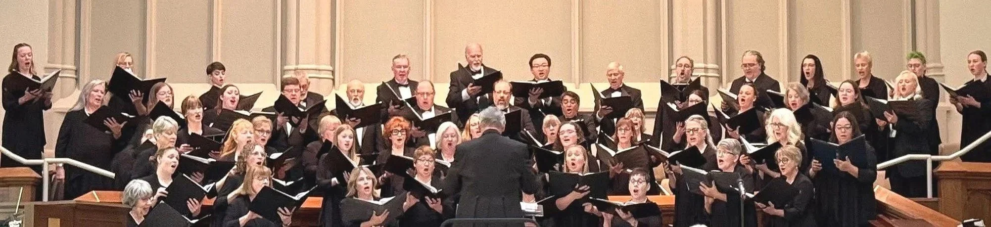 May 2026 photograph of subset of 50 singers in concert attire holding sheet music during a performance with Steve Eubank directing.