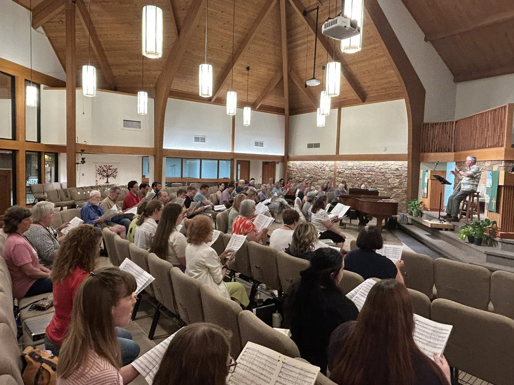 Photograph of 70 seated singers with sheet music at a rehearsal in a church, with Steve Eubank directing.