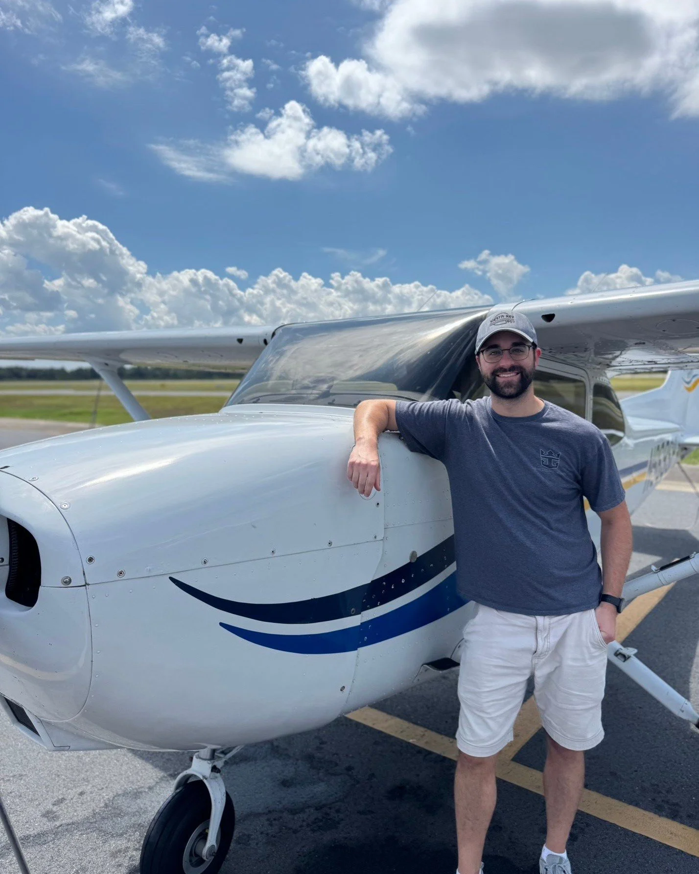 A man wearing a gray cap, glasses, a dark gray T-shirt, and white shorts standing next to a small white airplane with blue and black accents on a sunny day with a partly cloudy sky.
