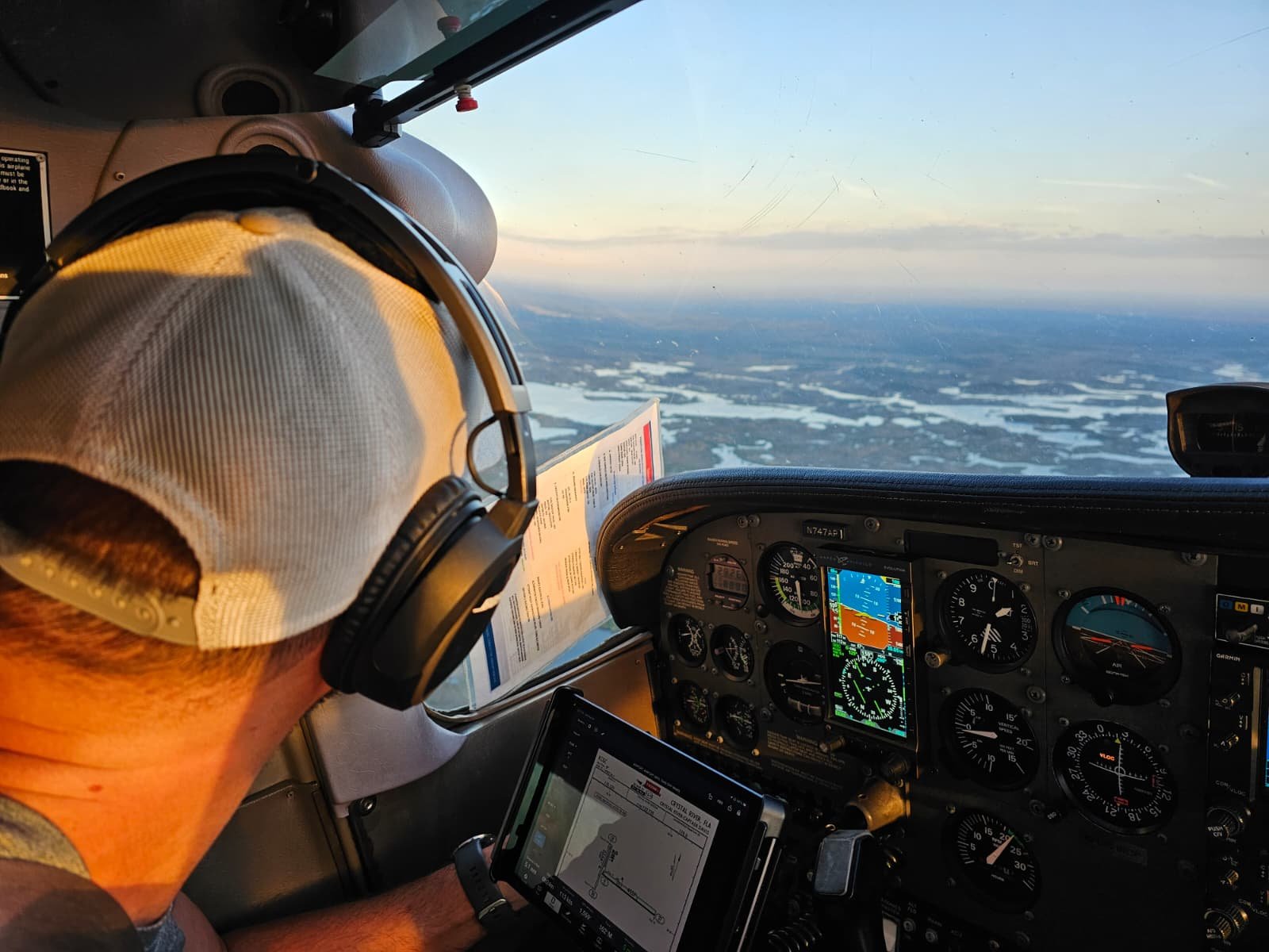 A pilot wearing a white cap and headphones flying an airplane over a landscape with water and land at sunset.