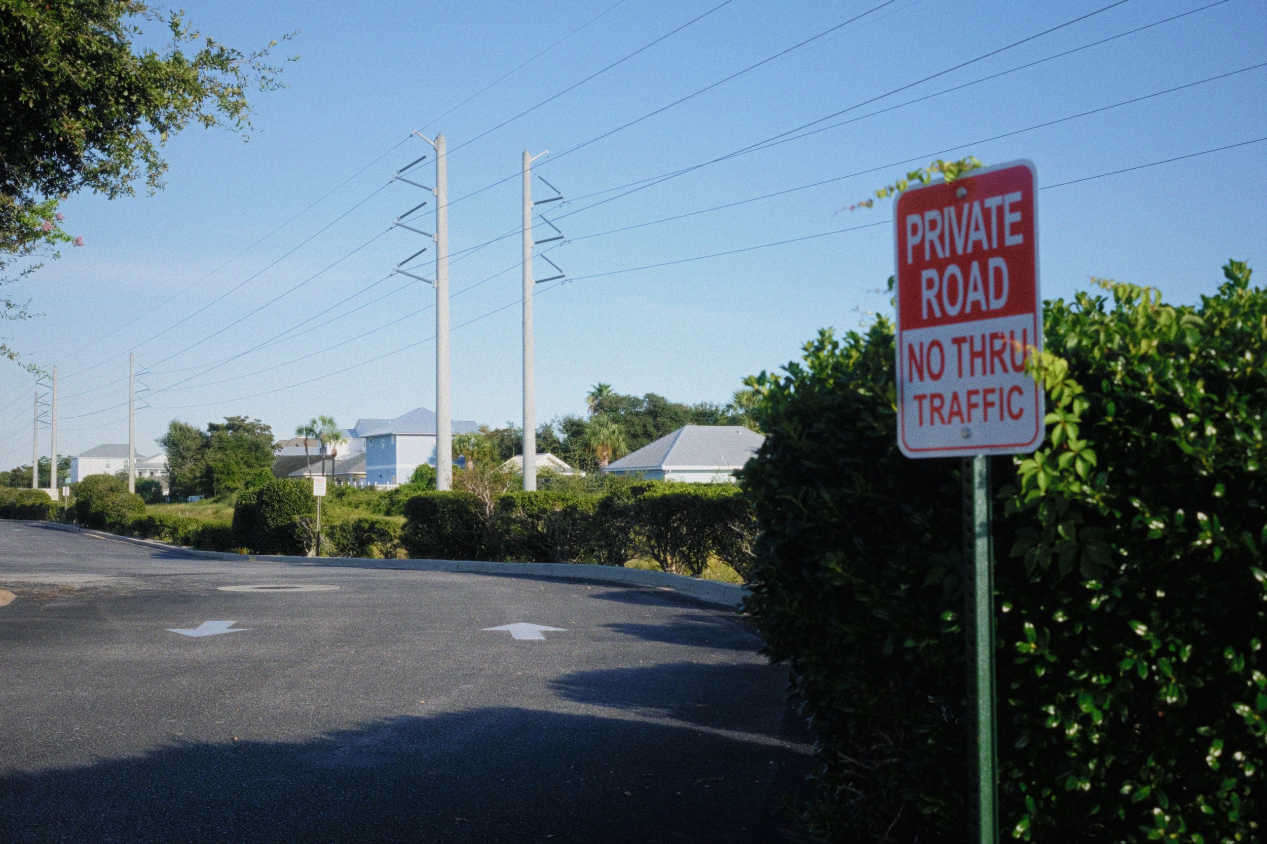 A red and white sign reading 'PRIVATE ROAD NO THRU TRAFFIC' in front of green bushes, with power lines and houses in the background on a clear sunny day.