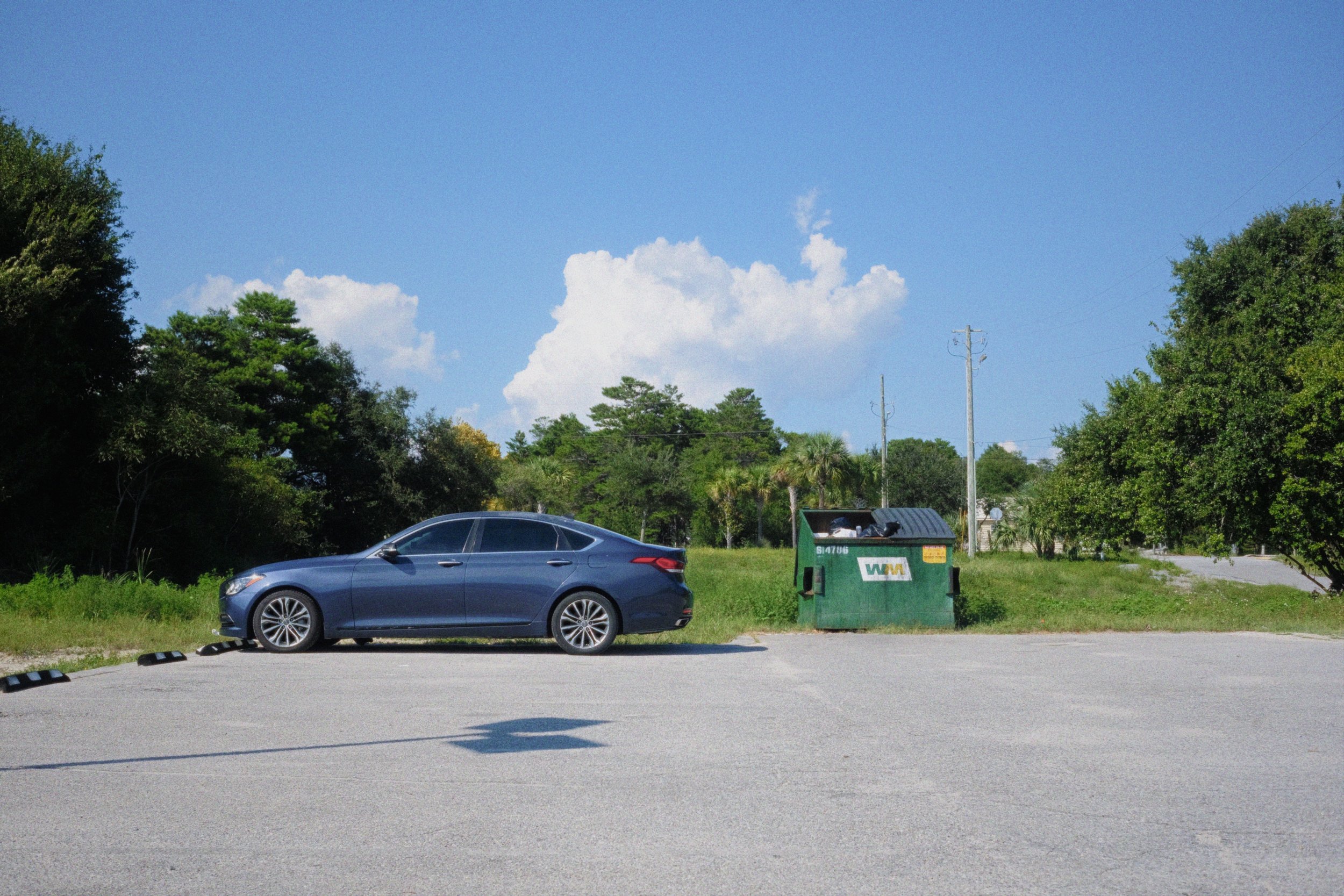 A blue sedan car parked on a paved lot with green and black wheel stops, a green trash bin, and trees and power lines in the background under a blue sky with some white clouds.