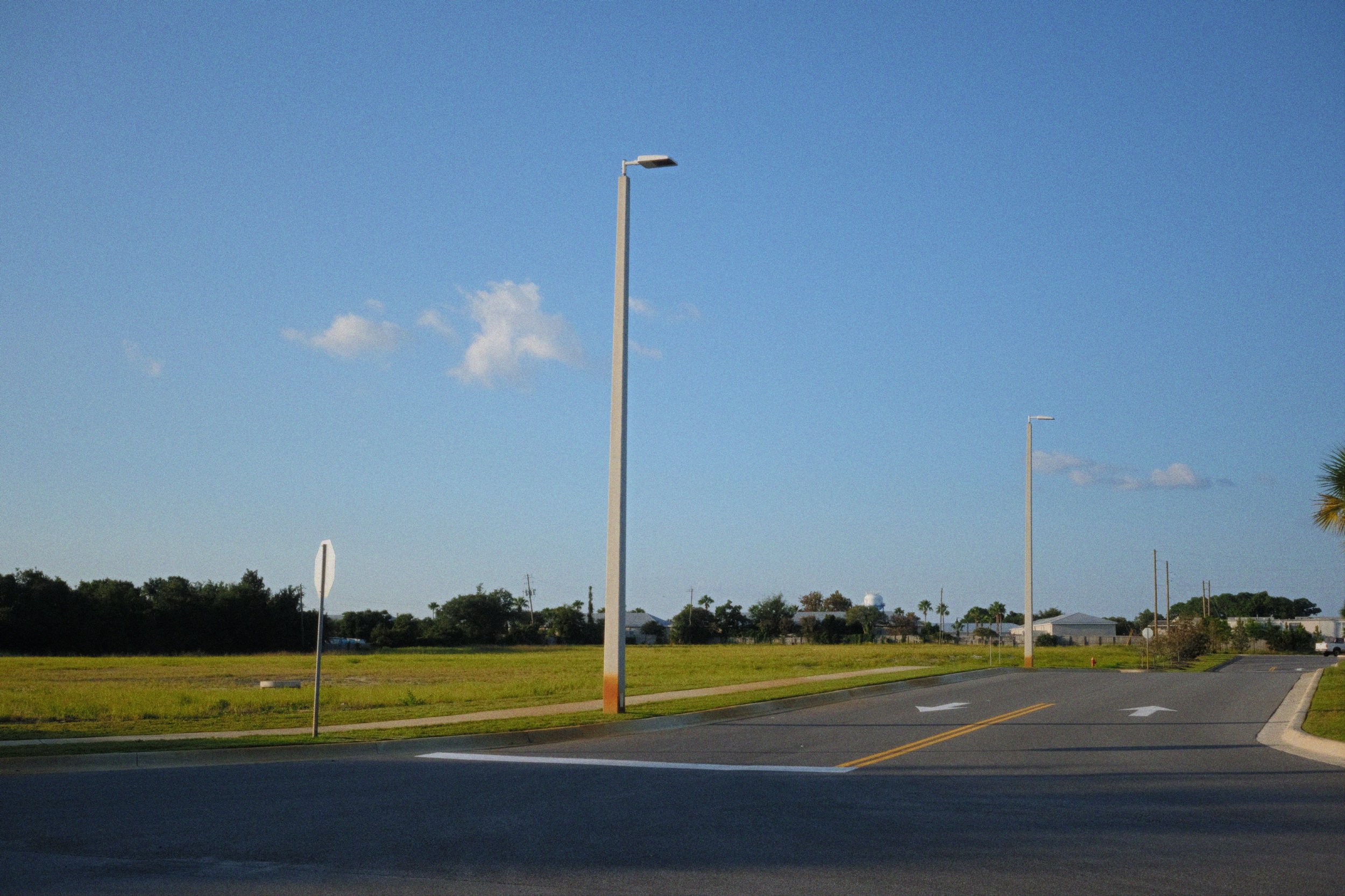 A suburban street scene with streetlights, a stop sign, a grassy area, and houses in the background under a bright blue sky with a few clouds.