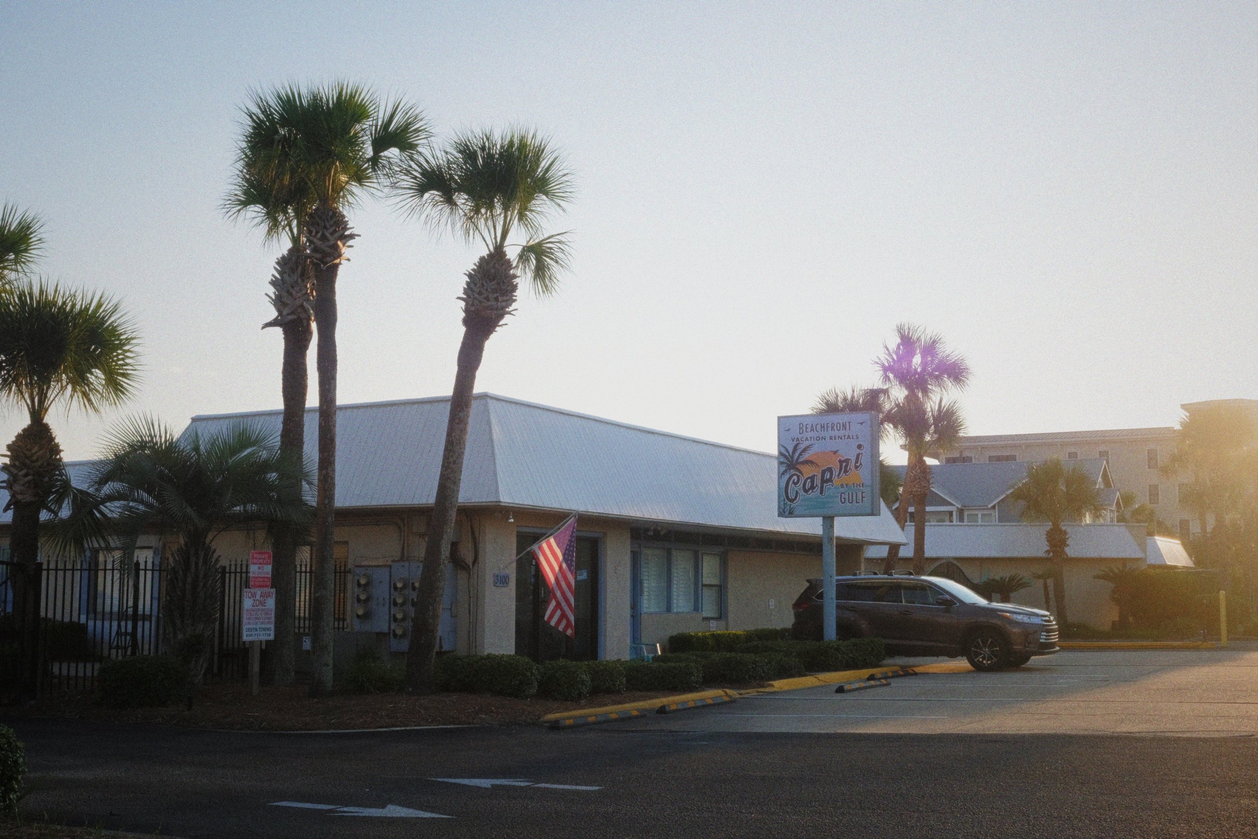 A beachfront building with a sign that says 'Capri by the Gulf' surrounded by palm trees. There is a parking lot with a black car and an American flag in front.