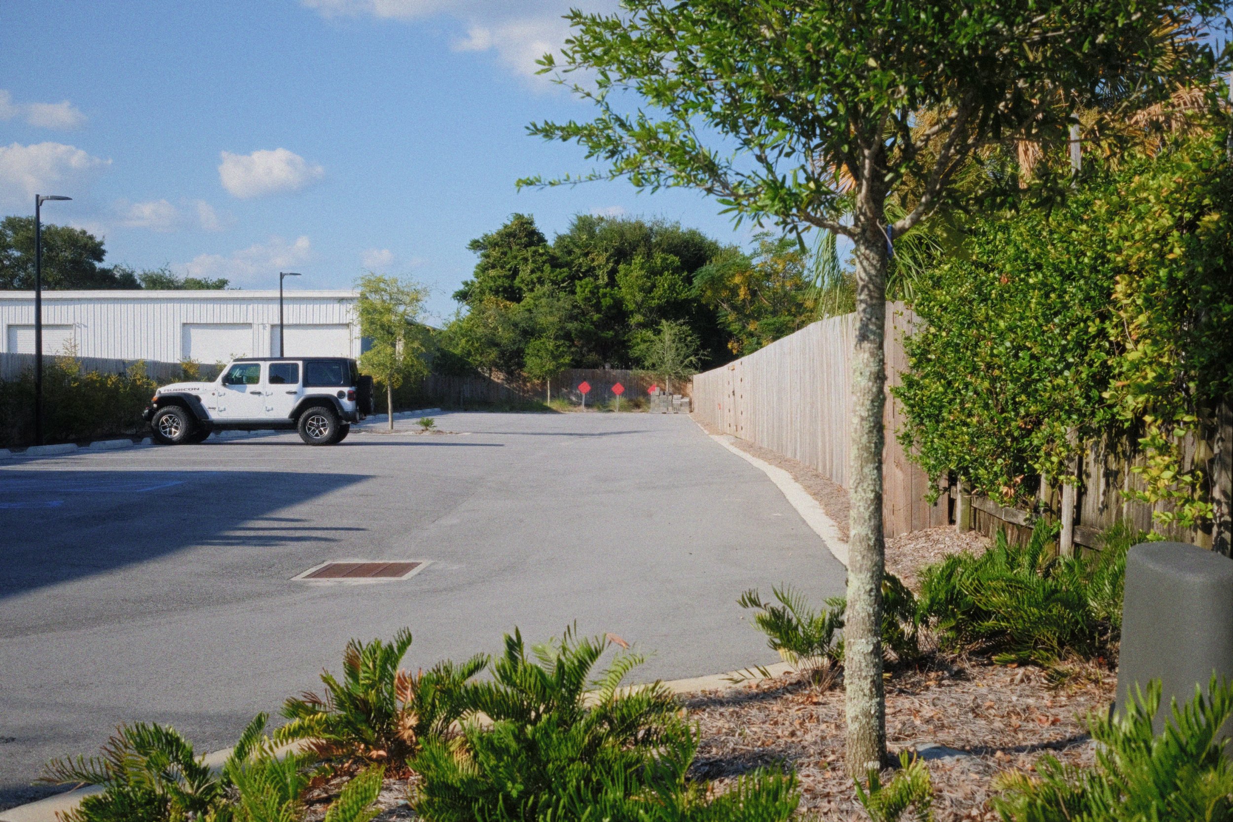 A parking lot with a white Jeep Renegade parked near a wooden fence and lush green trees, under a bright blue sky with a few clouds.