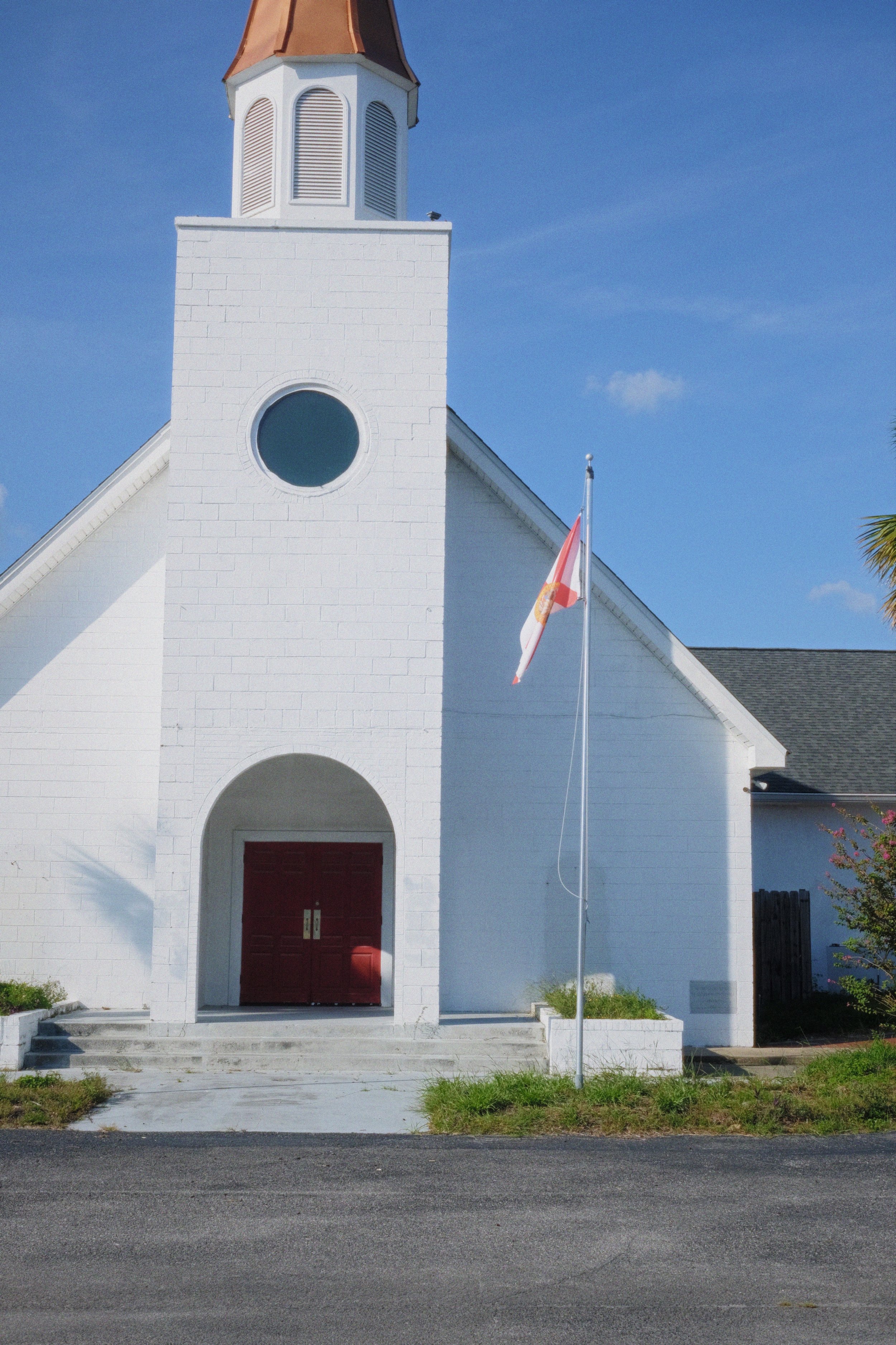 A white church with a small steeple topped with a copper-colored roof, a circular window, red double doors, a flagpole with a red and white flag, and steps leading to the entrance, under a clear blue sky.