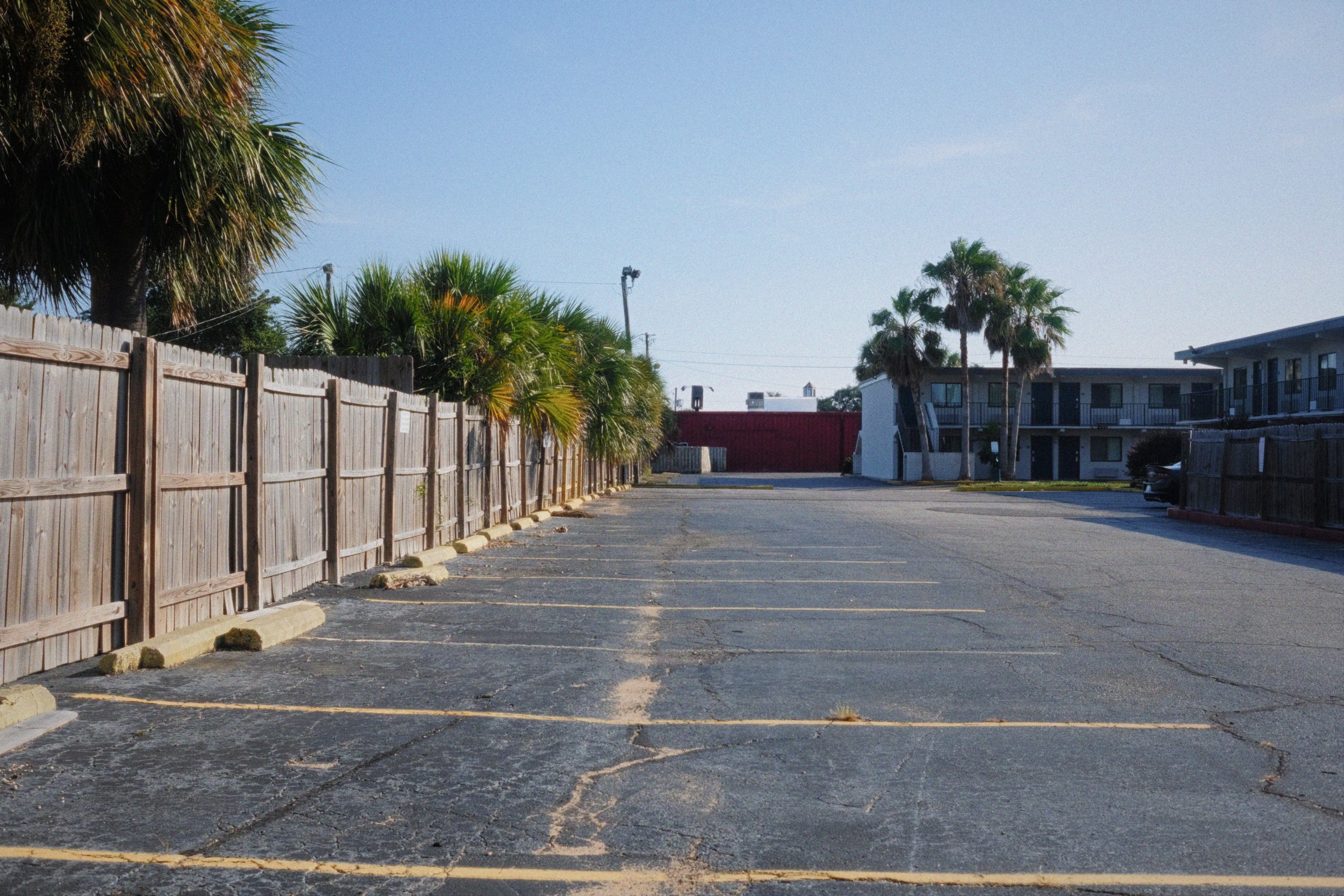 A nearly empty parking lot with yellow parking lines, a wooden fence on the left, and a building with balconies on the right. There are palm trees and a clear blue sky.