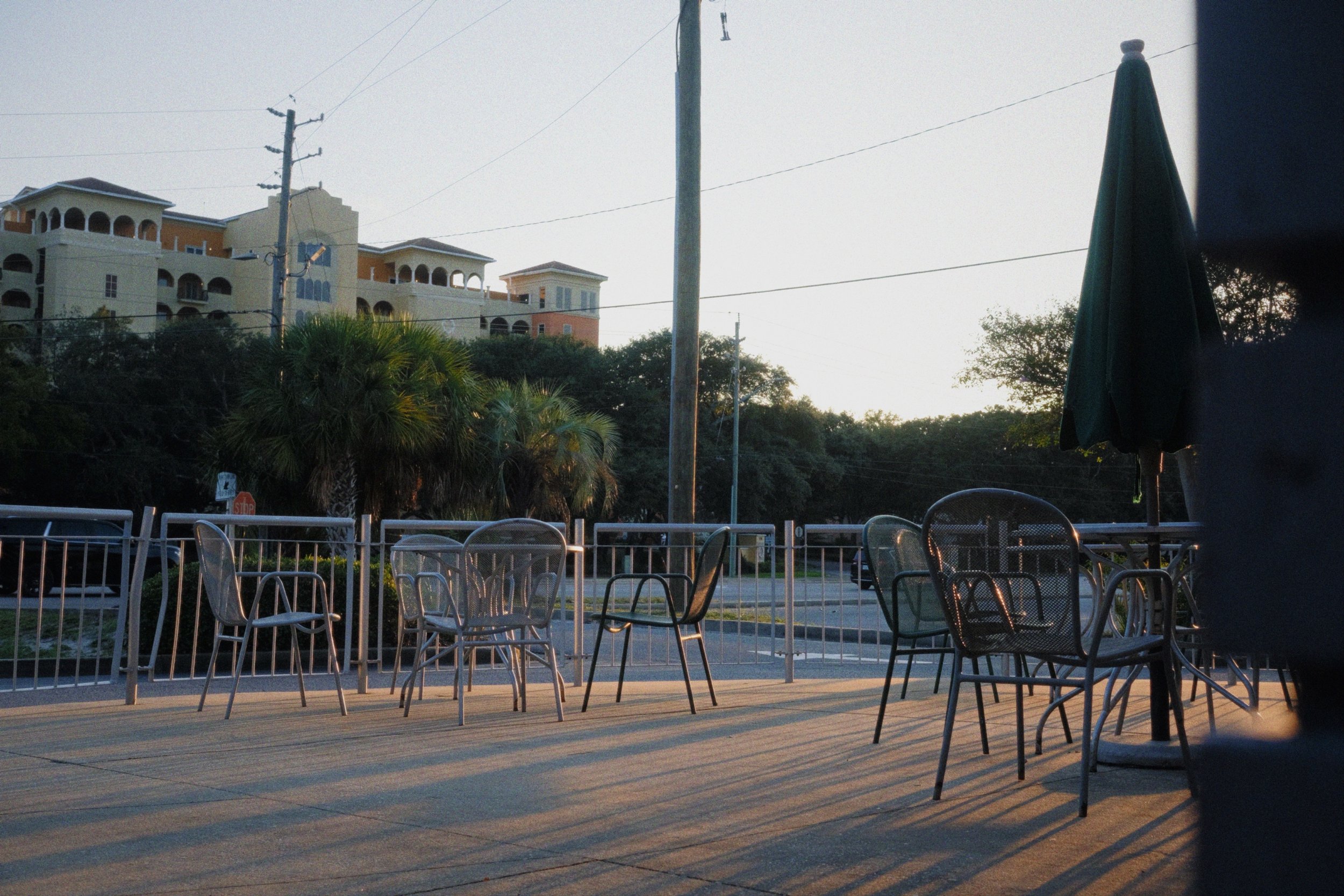 An outdoor patio with metal chairs and tables, some with umbrellas, during sunset. In the background, there are lush trees and a large, multi-story building with balconies and arched windows. Power poles and wires are visible, and the scene has a tra