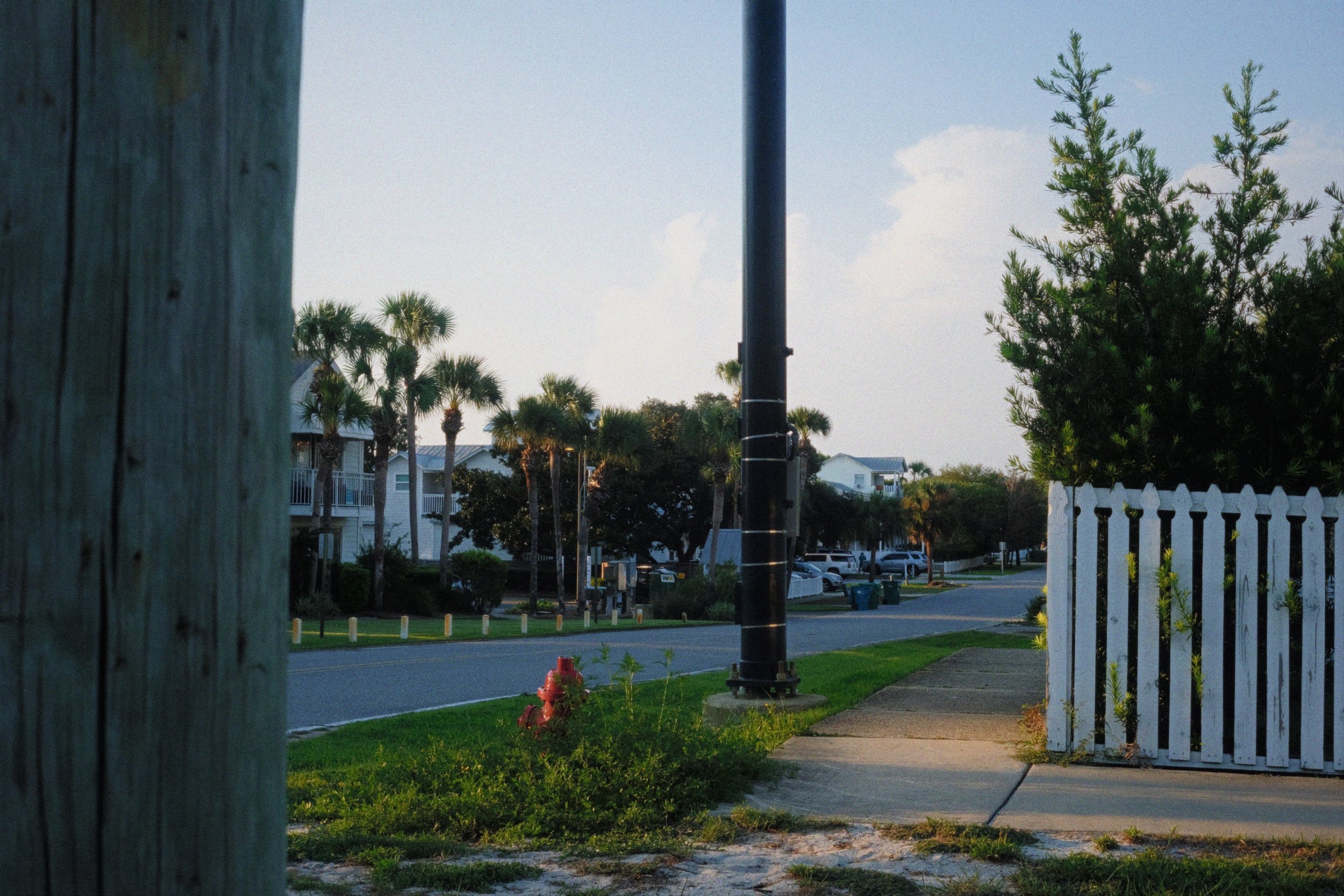 A suburban neighborhood street scene during daytime with clear skies, trees, houses, cars, a white picket fence, a utility pole, and a fire hydrant in the foreground.