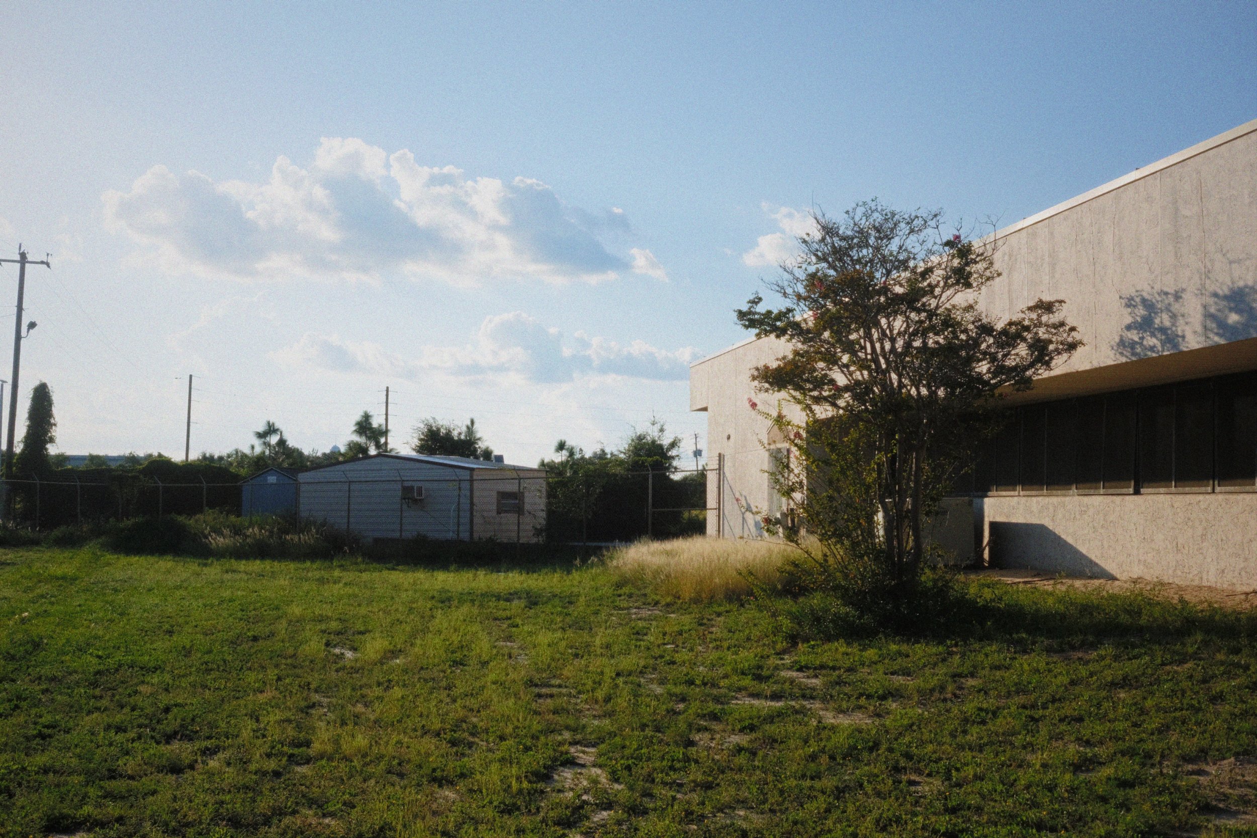A grassy area with a small tree, a large beige building, a chain-link fence, and utility poles against a blue sky with scattered clouds.