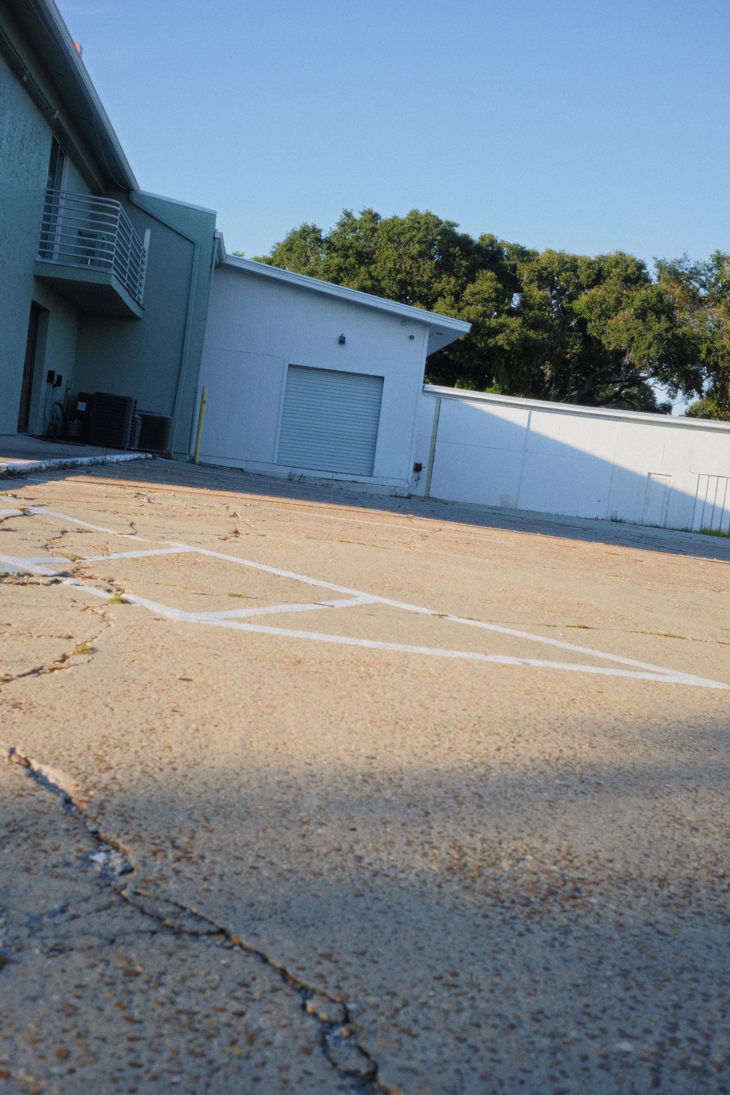 An empty parking lot with visible cracks on the pavement, adjacent to a light-colored building with a roll-up garage door and air conditioning units, and a background of green leafy trees under a clear blue sky.