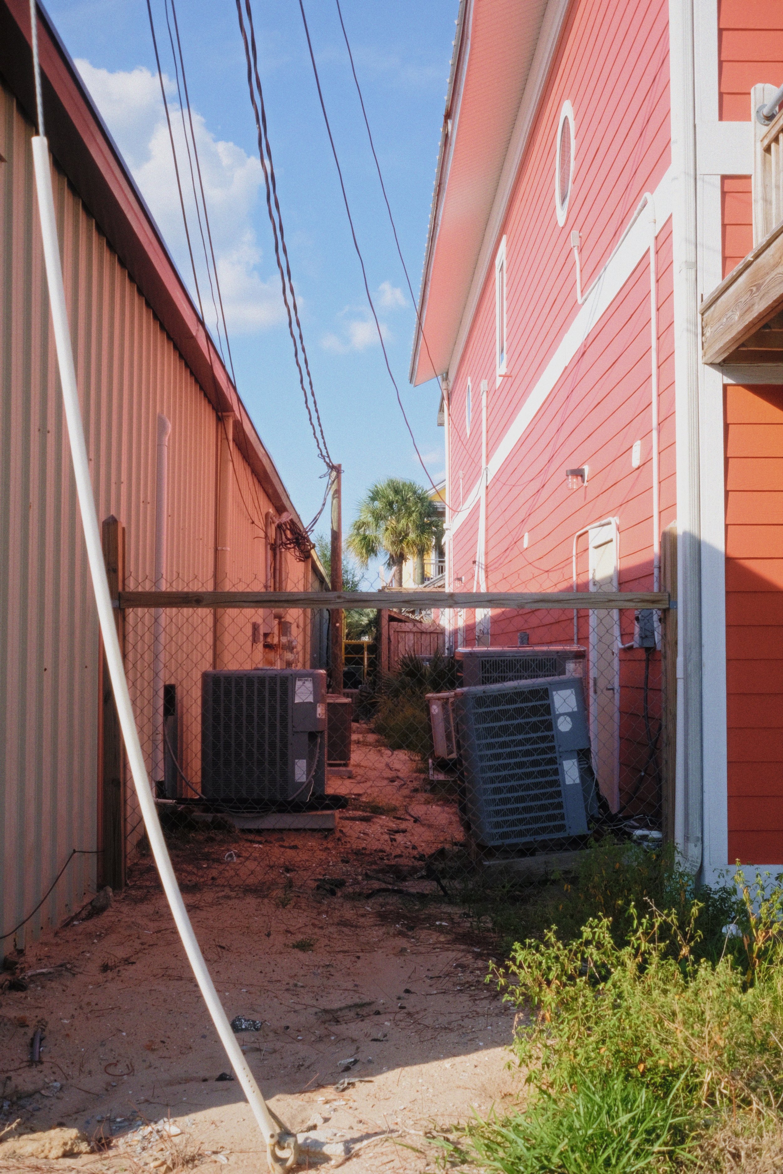 Alleyway between a pinkish-red building and a beige building with air conditioning units and electrical wires, with some plants and dirt on the ground.